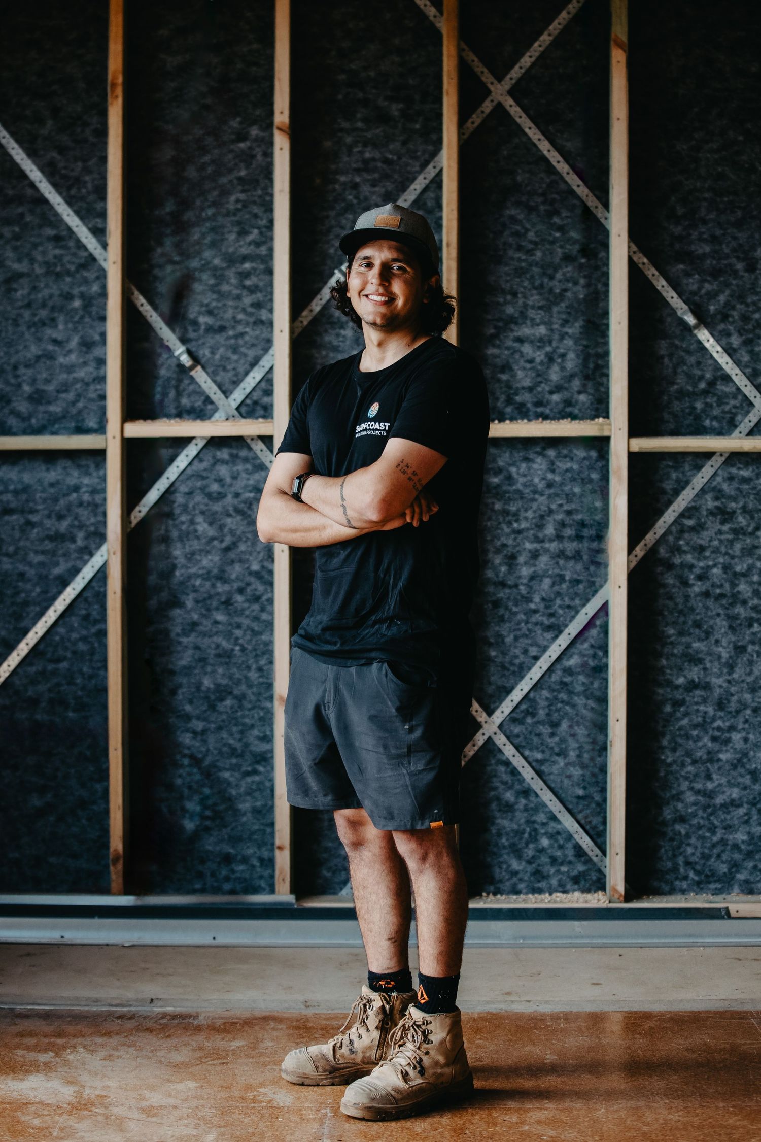 Young tradesman in black t-shirt, shorts, and work boots stands confidently with arms crossed in an unfinished building interior.