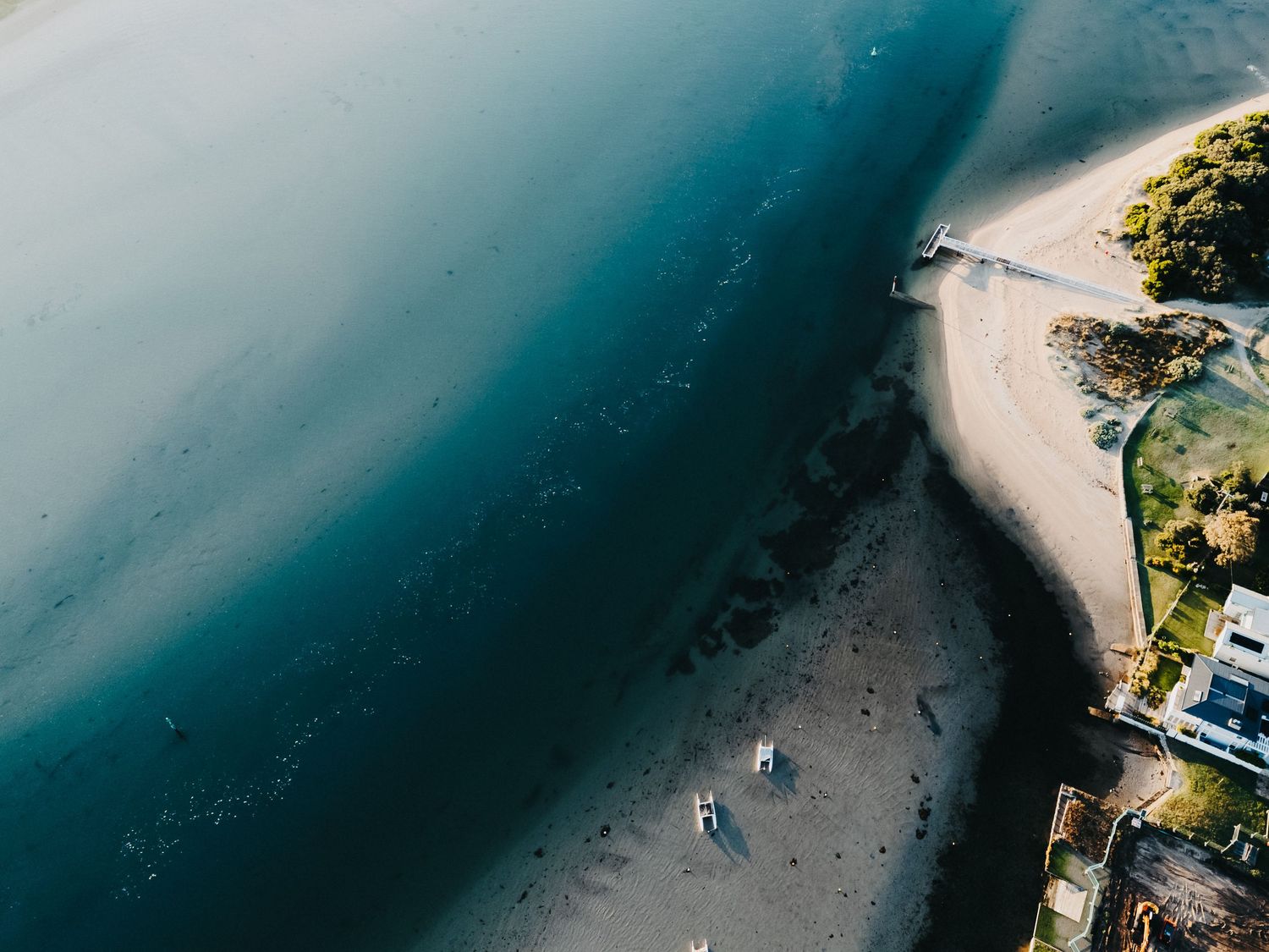 Aerial drone view of a sandy beach coastline meeting calm teal ocean waters with a small pier nearby.