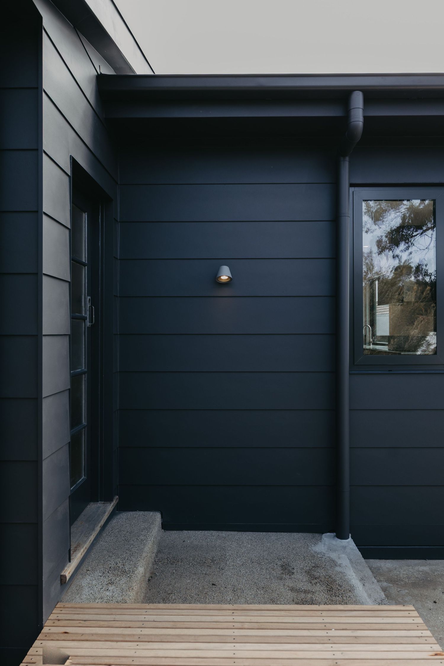 Exterior of contemporary dark navy cladded home with wall-mounted light fixture, concrete steps, and timber deck.