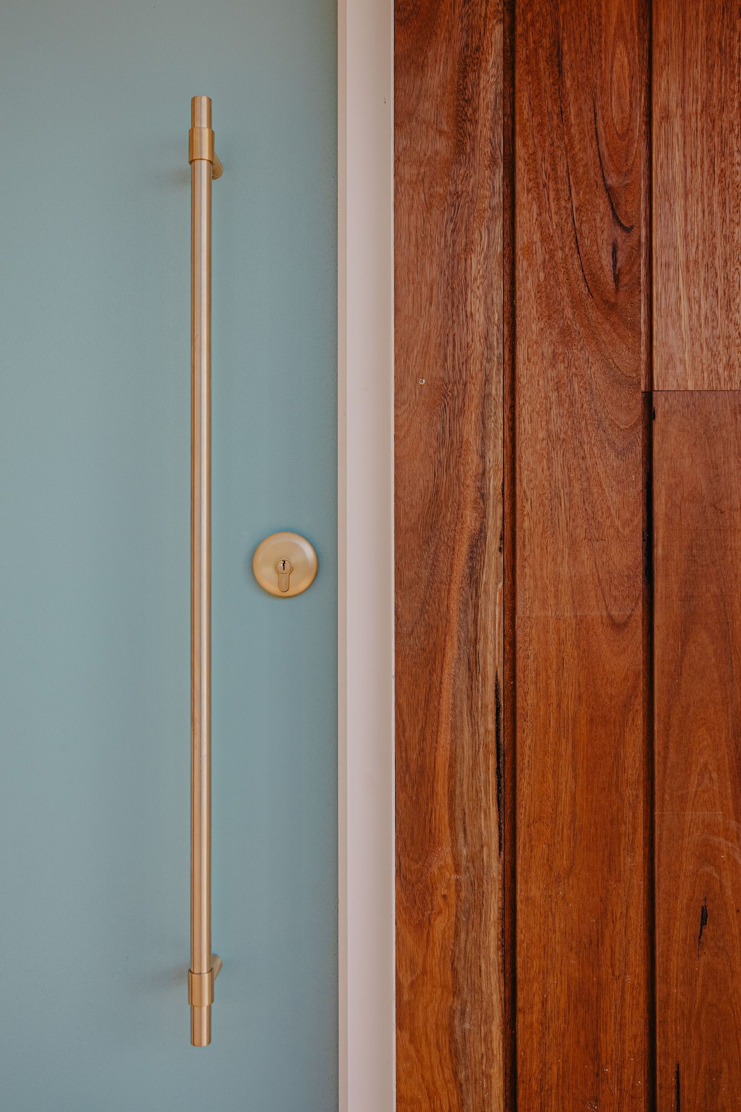 Close-up of a sleek brass pull handle and round knob on a teal door beside rich timber wood cladding.