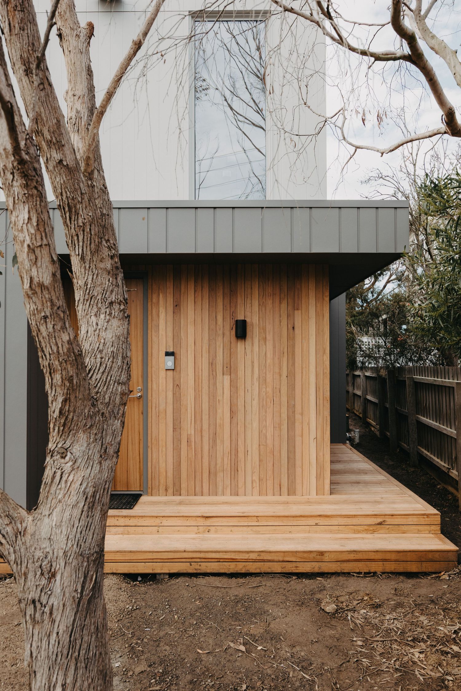 Modern home entrance with cedar timber cladding, wooden deck steps, and intercom panel framed by a bare tree.