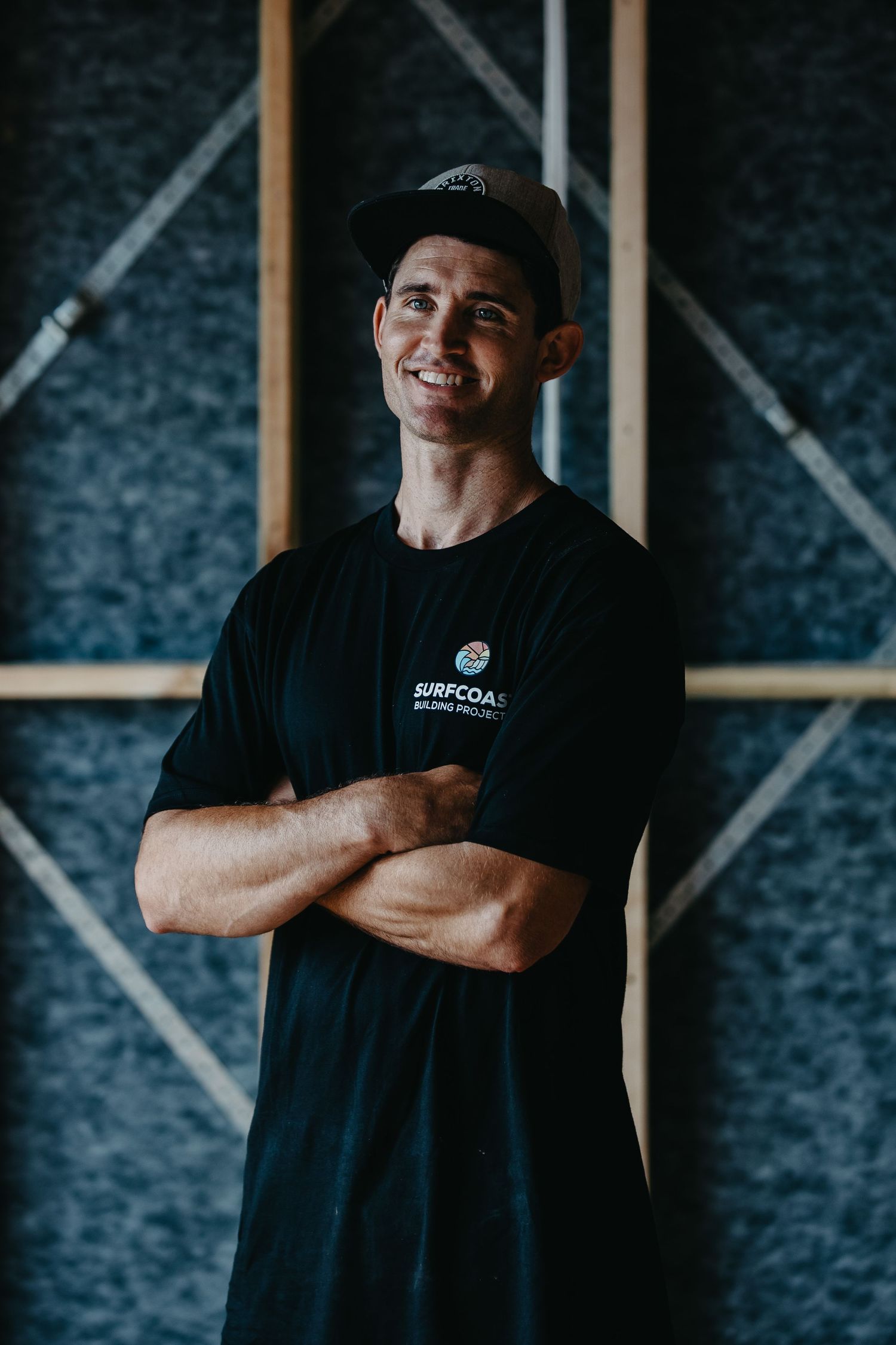 Smiling athletic man in black Surfcoast t-shirt and cap stands with arms crossed against geometric wall.