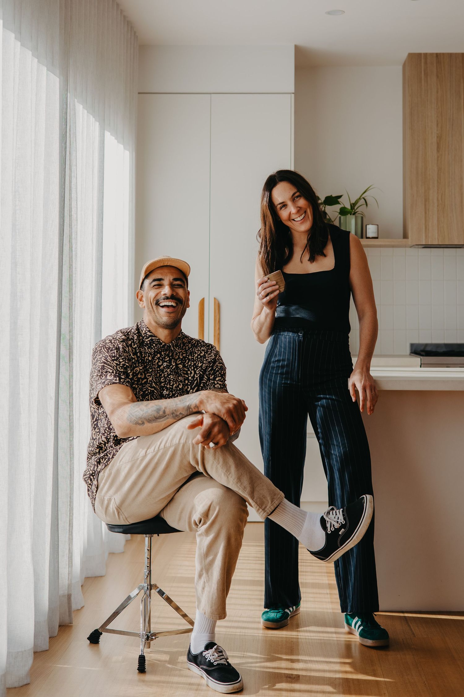 A smiling man and woman pose casually in a modern apartment, he seated on a stool, she standing beside him.