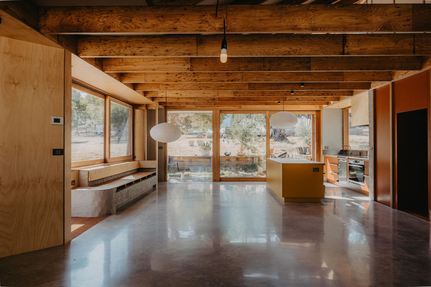 Open-plan interior with exposed timber ceiling beams, polished concrete floors, plywood walls and large windows framing bushland views.