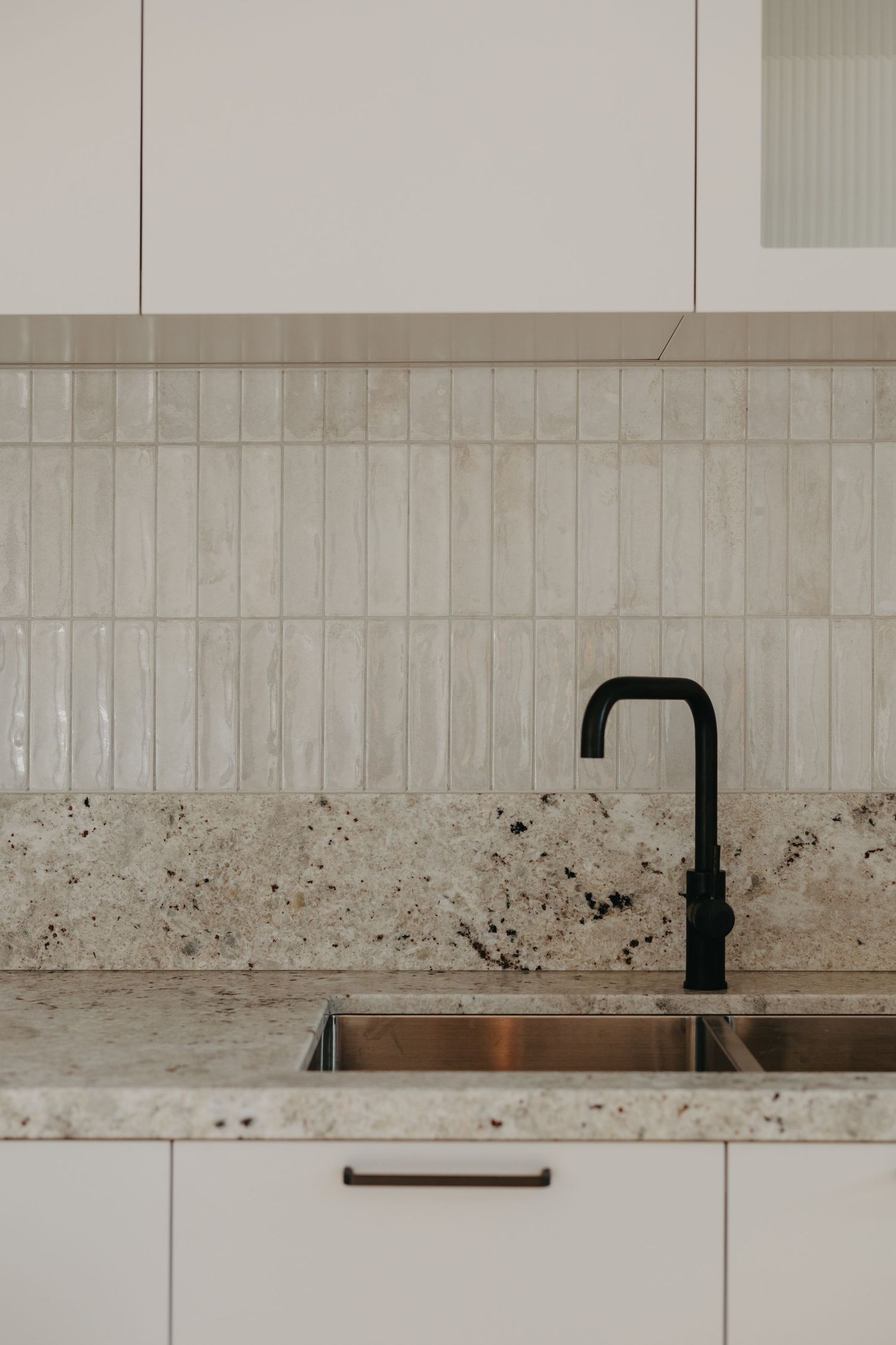 Modern kitchen sink with black matte faucet, granite countertop, white cabinets, and vertical ceramic tile backsplash.