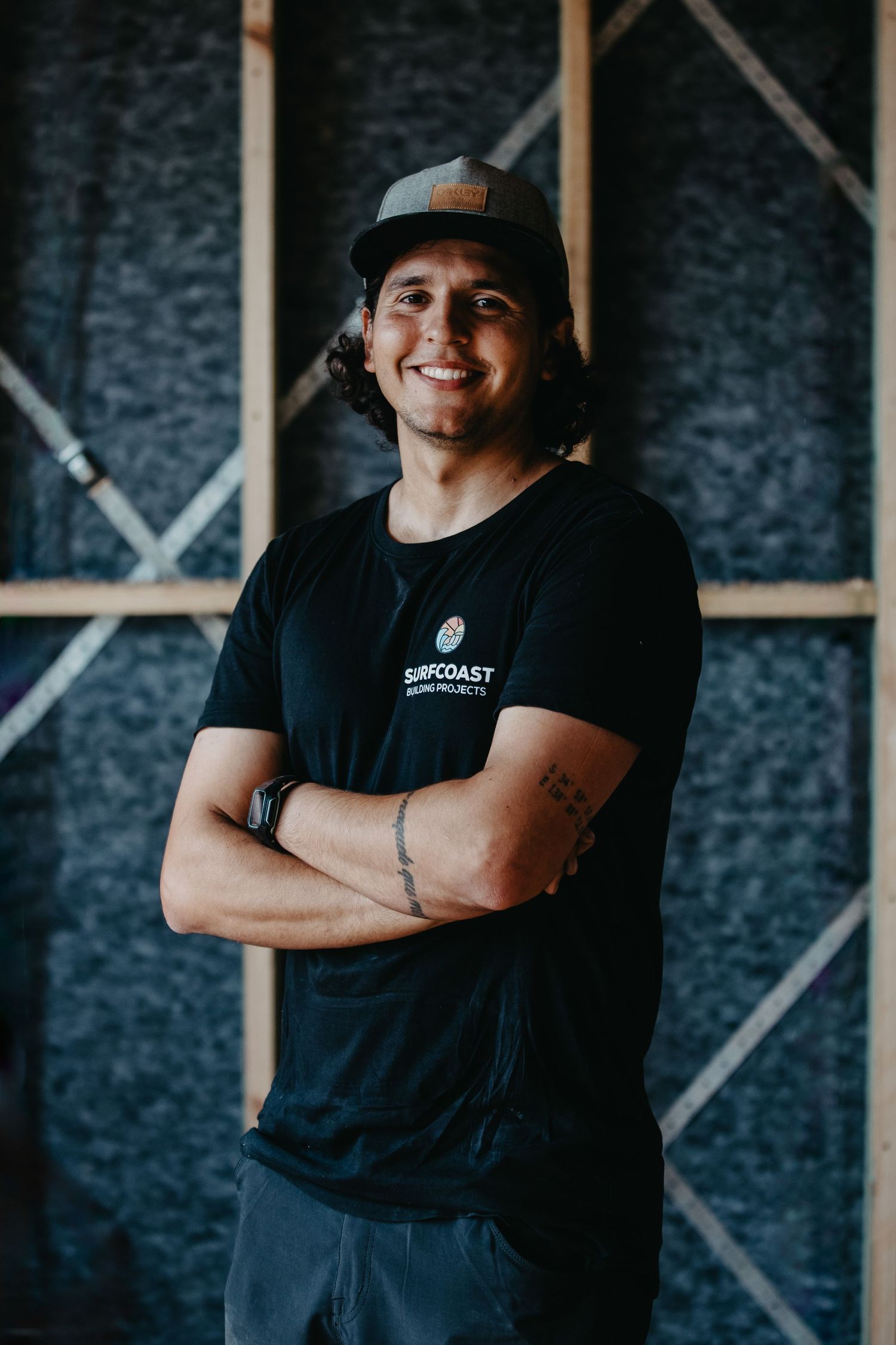 Young man in black Surfcoast t-shirt and cap smiling with arms crossed in a construction site.