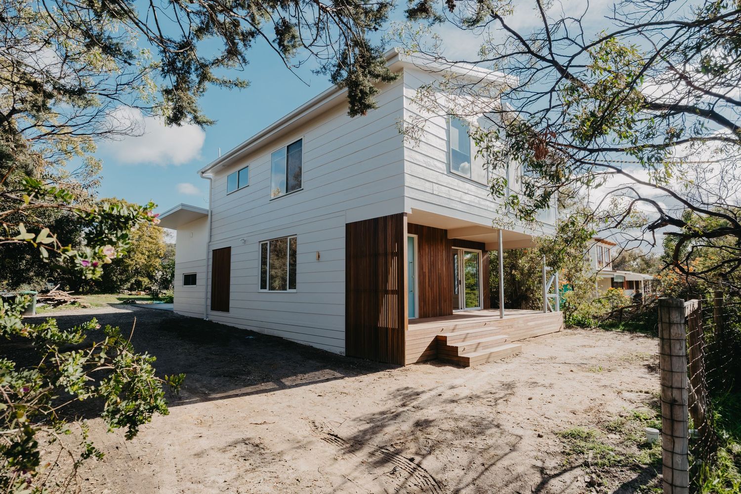 Two-storey white weatherboard house with timber deck entry, surrounded by native trees and gravel driveway.
