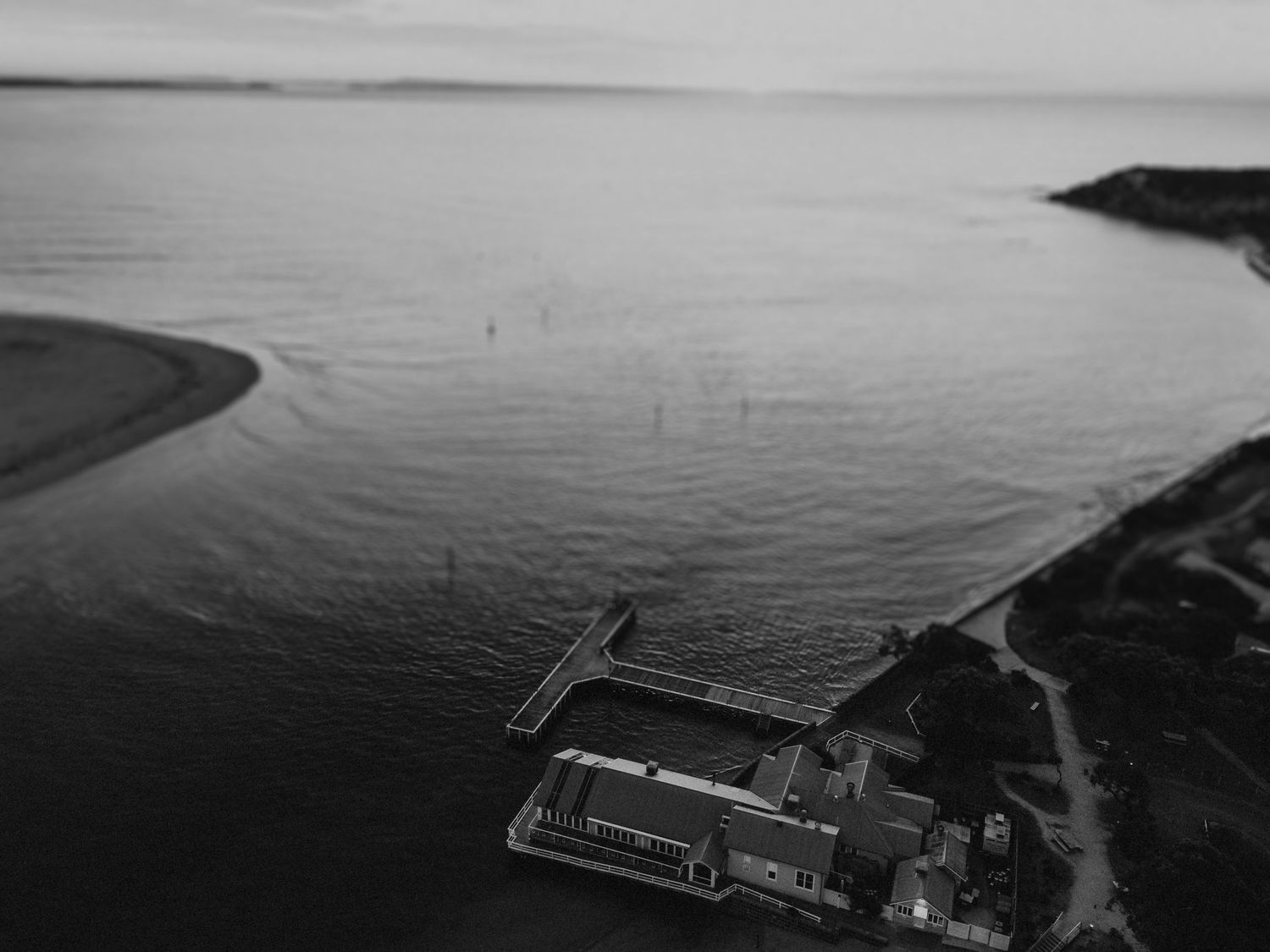 Aerial black and white photo of a coastal building with a pier extending into calm ocean waters at dusk.