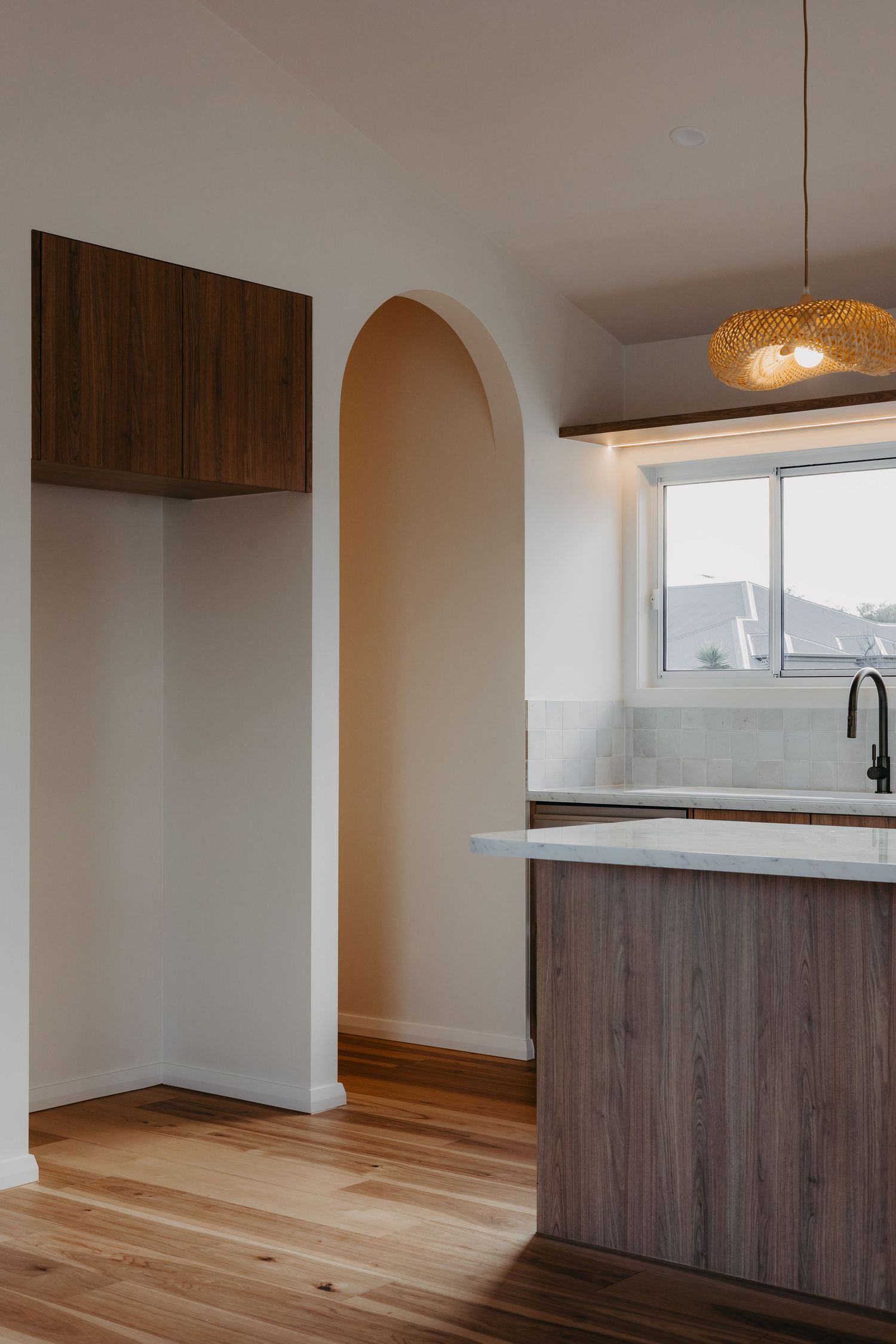 Kitchen entry with dark timber wall cabinet, arched pantry doorway, marble island bench, pendant light, and wide plank timber floors.