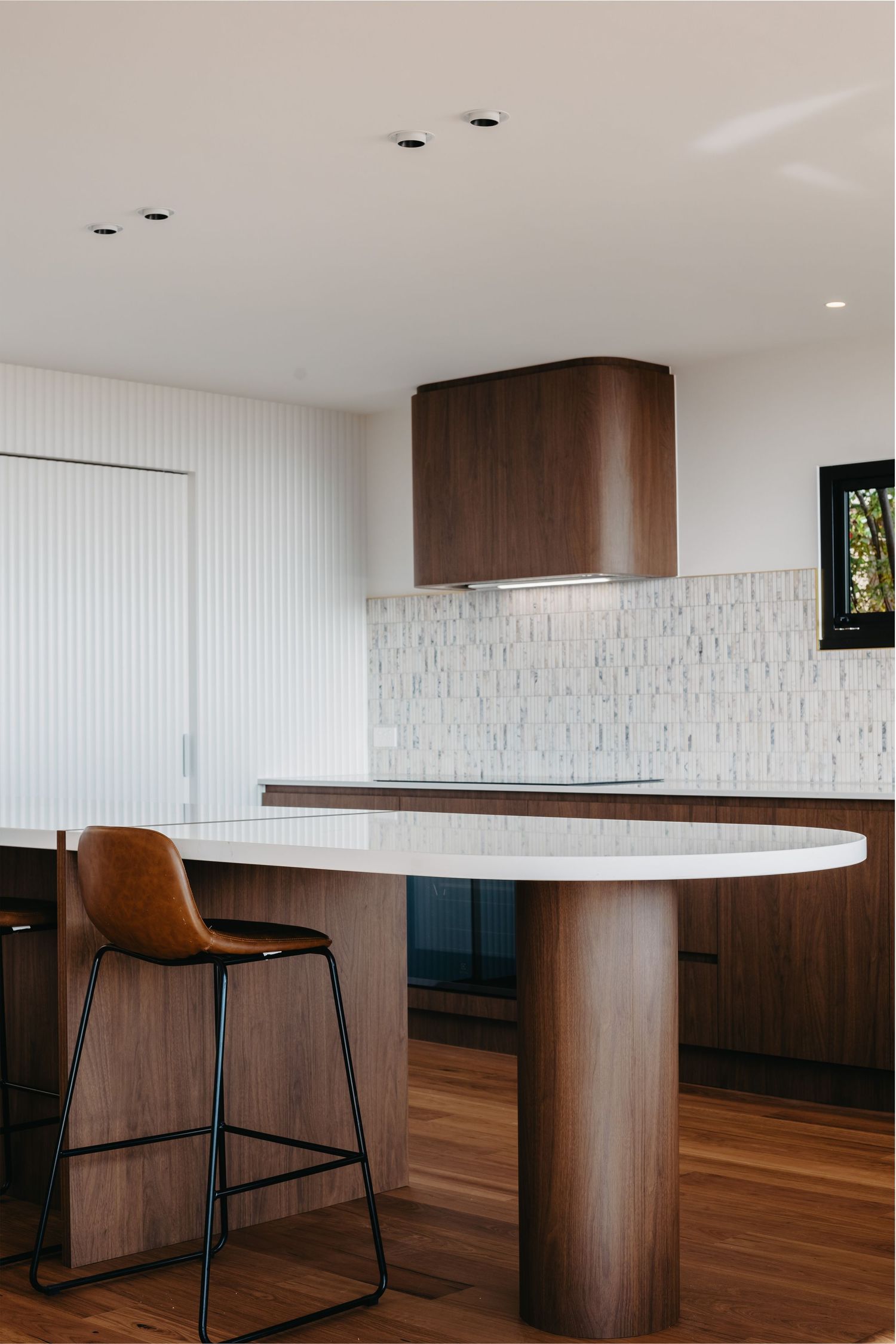Modern kitchen with curved timber island, white countertop, walnut cabinetry, and leather bar stool seating.