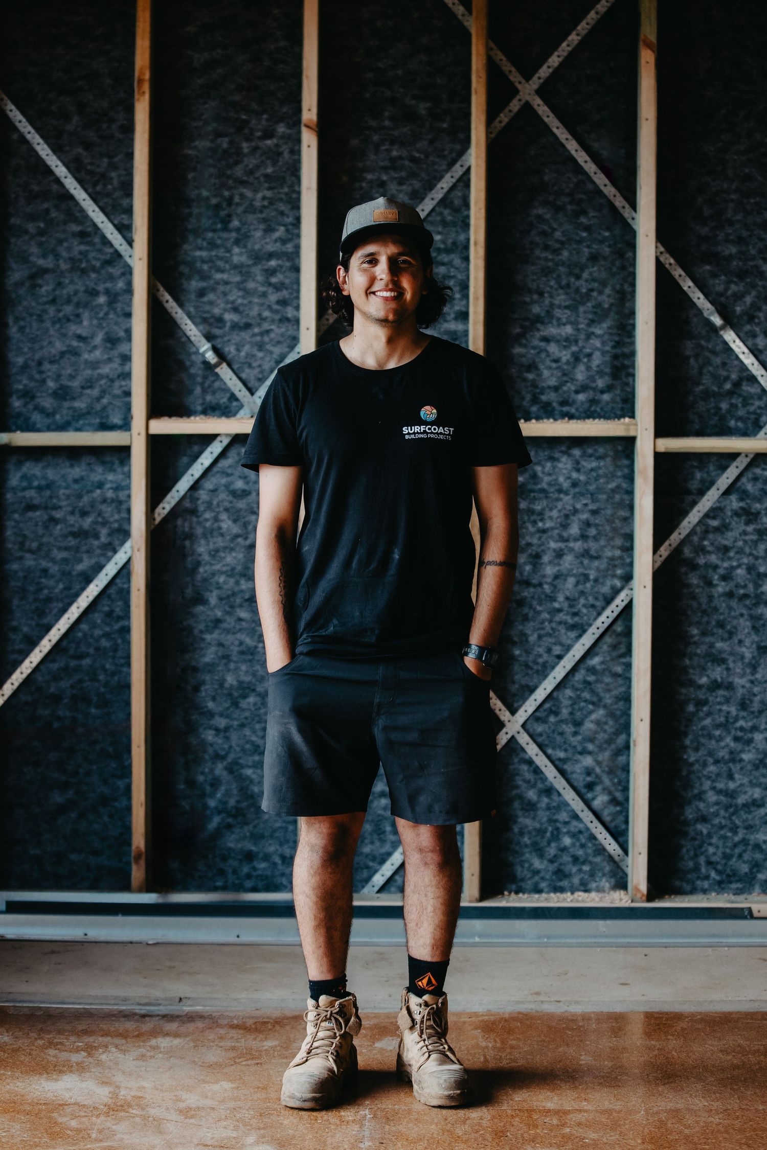 Smiling young man in black t-shirt, dark shorts, and work boots standing in a construction site.