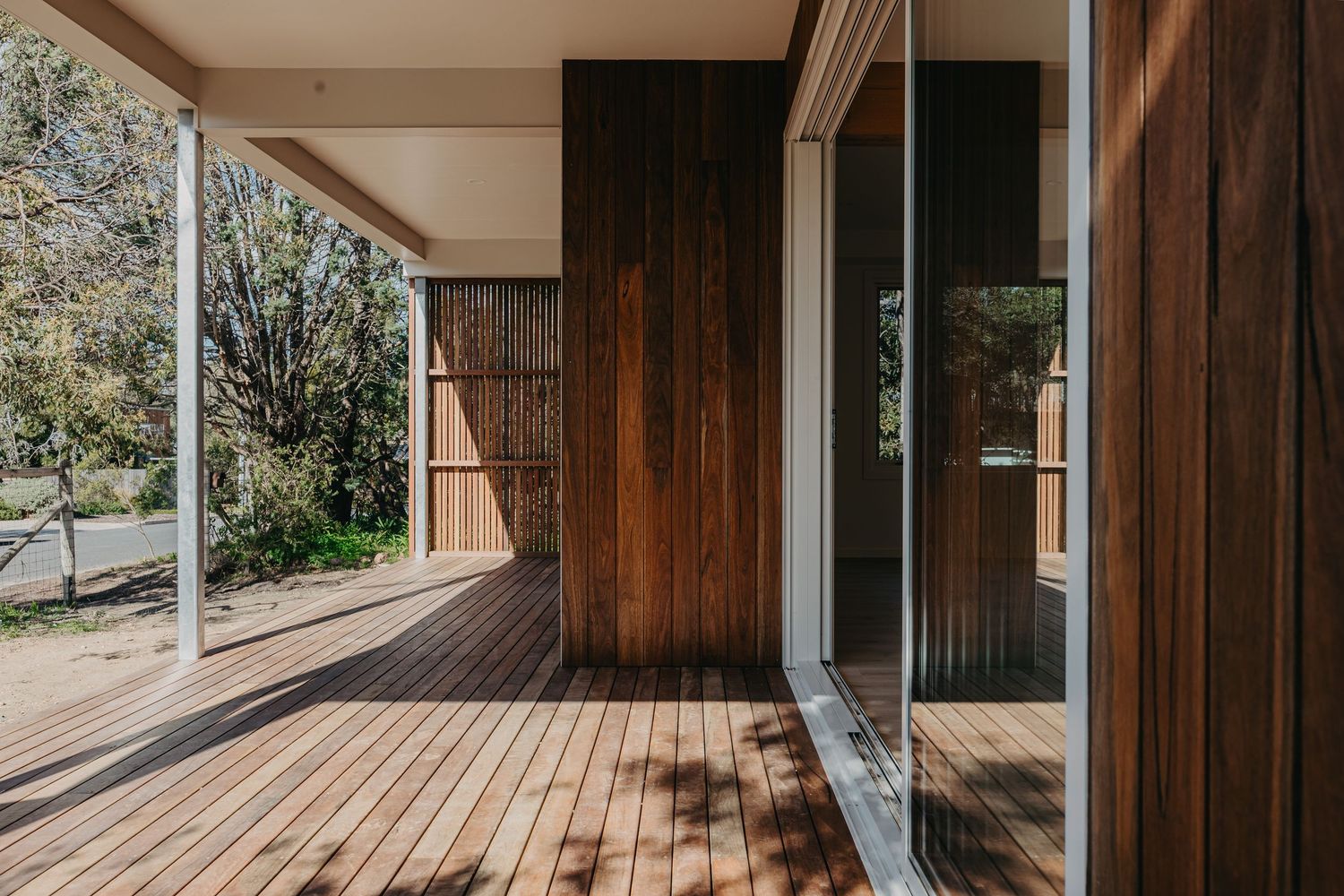 Covered timber deck with vertical wood feature wall, glass sliding doors, and surrounding native garden trees.