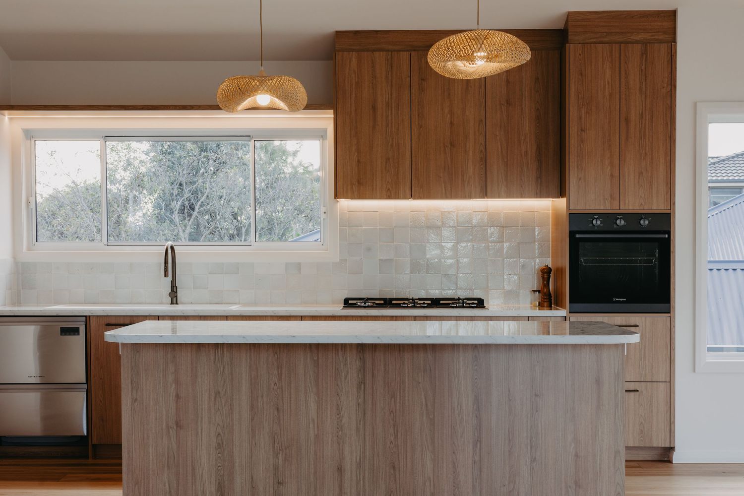 Modern kitchen with walnut cabinets, white marble island, gas cooktop, black oven, subway tile backsplash, and woven pendant lights.