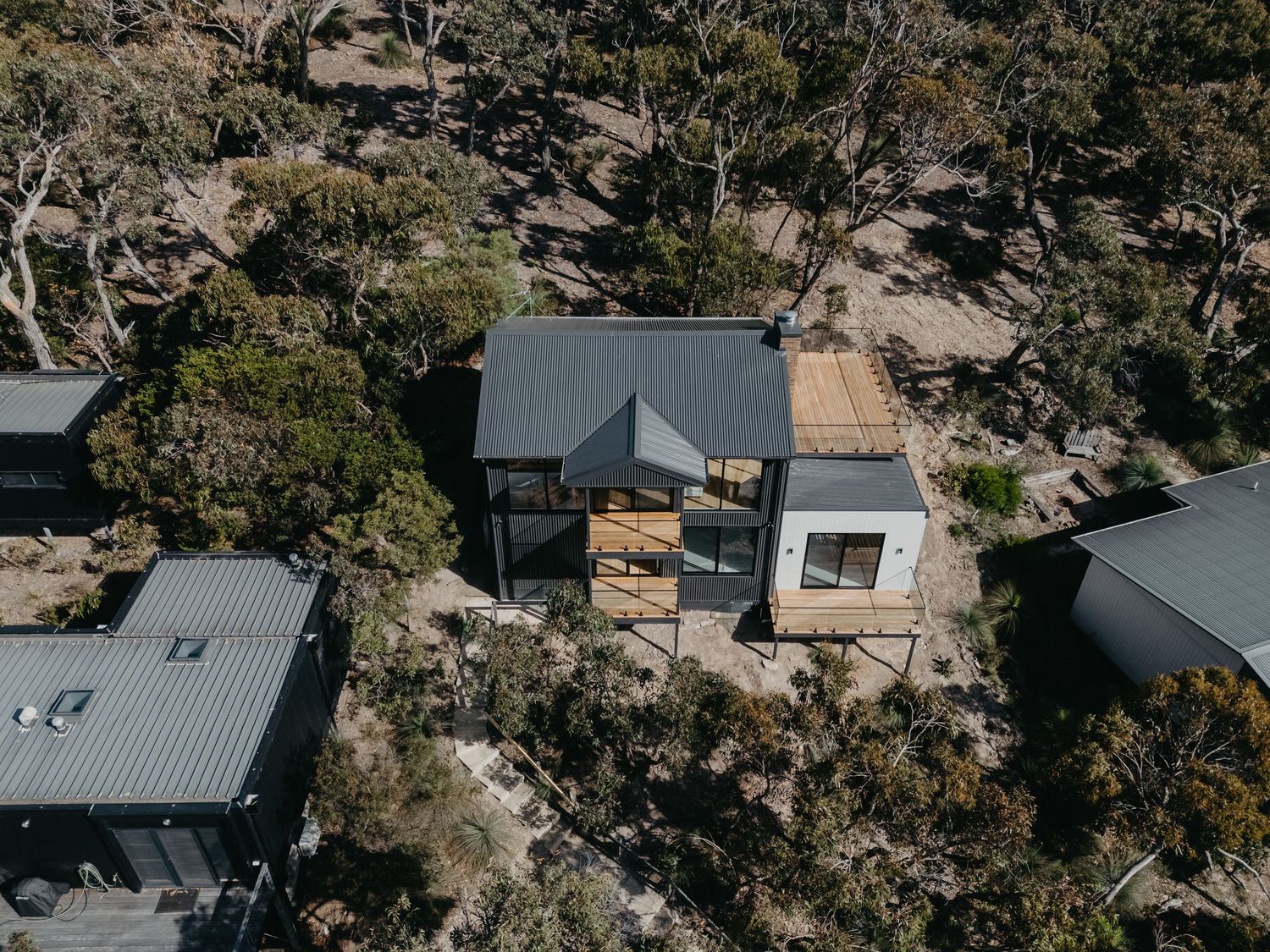 Aerial view of small modern cabin with timber deck surrounded by sparse bushland.