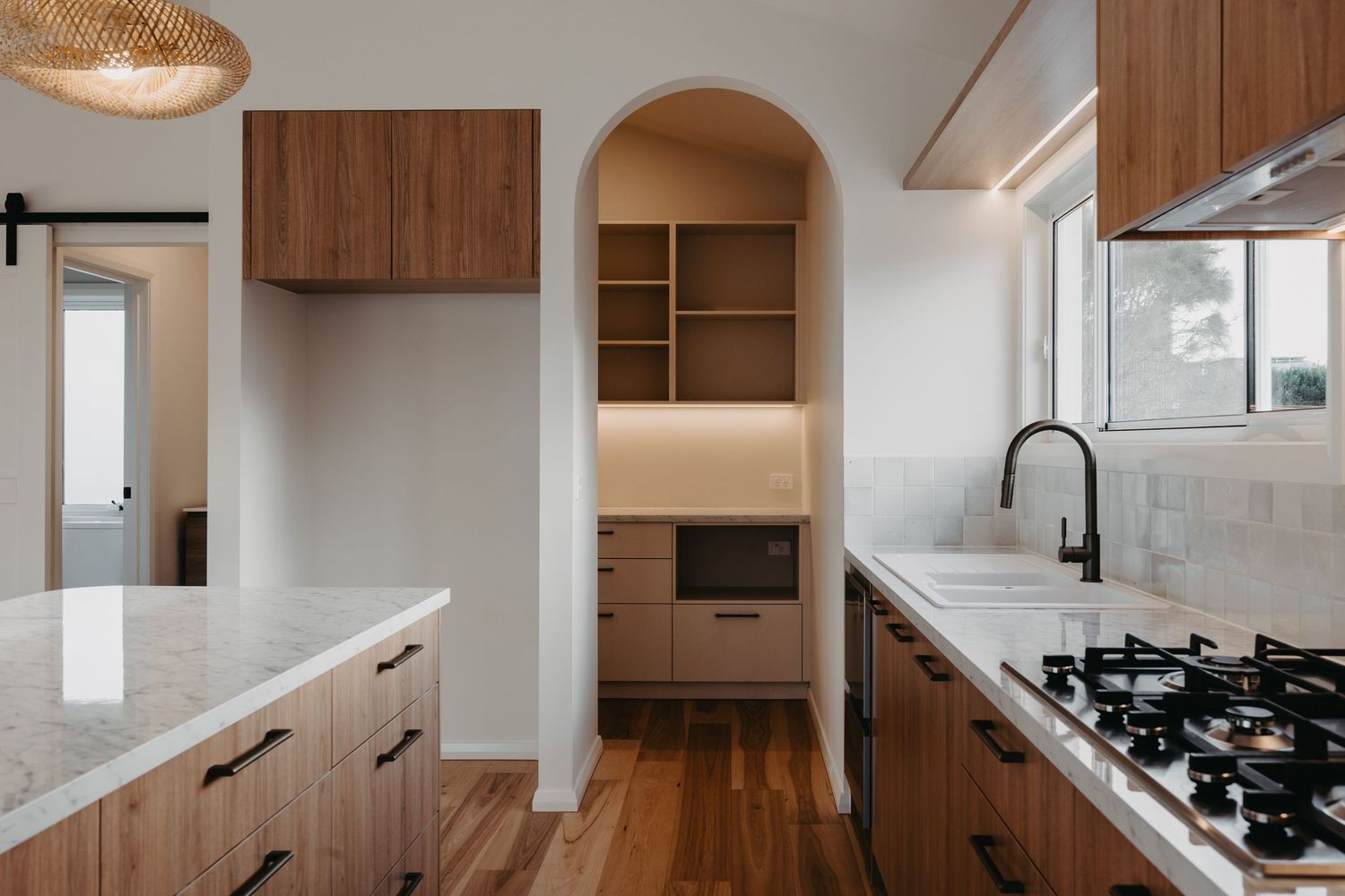 Modern kitchen with walnut cabinets, marble countertops, gas cooktop, and arched pantry doorway on hardwood floors.