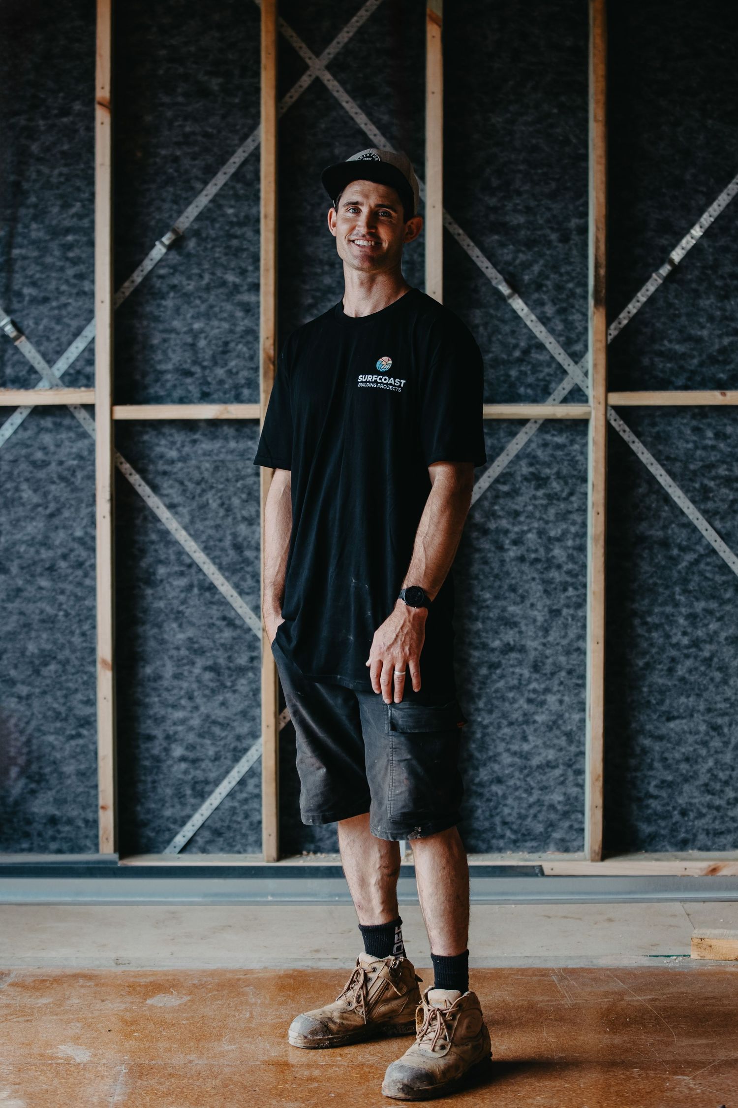 Man in black t-shirt, shorts and work boots standing in a timber-framed room.