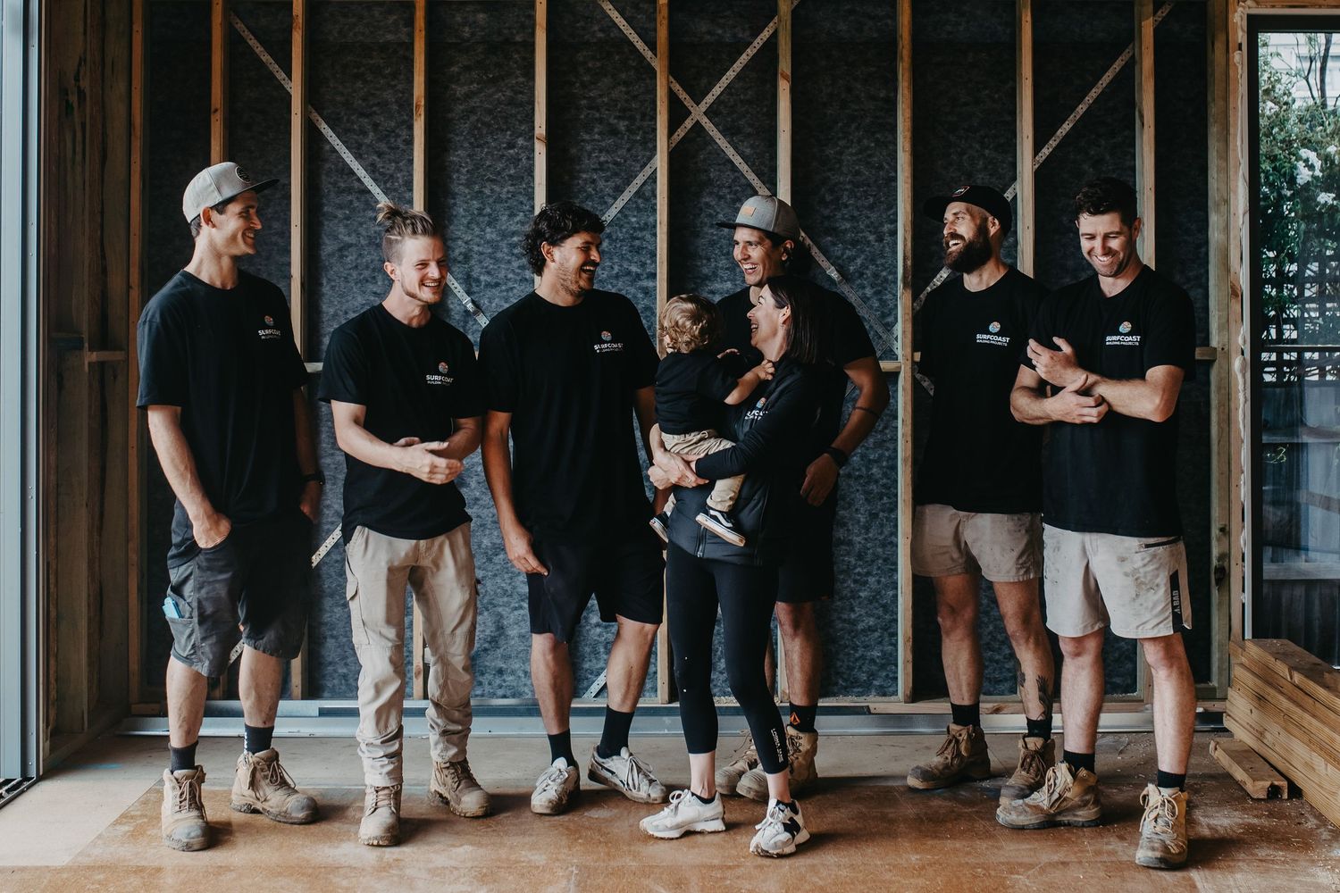 Seven construction workers in matching black shirts pose together inside a timber-framed building under renovation.