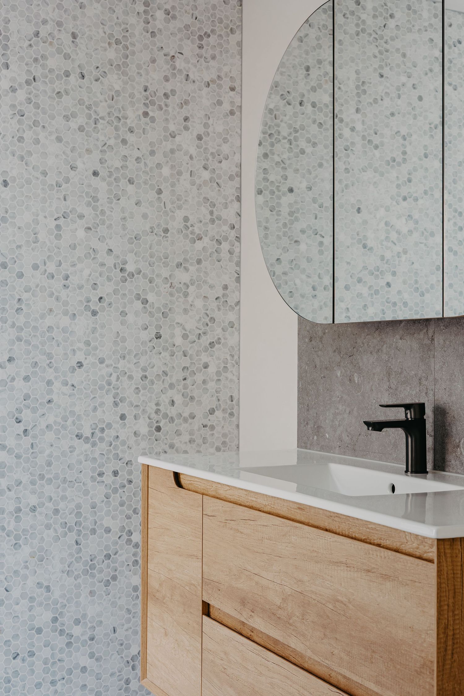 Modern bathroom vanity with timber cabinetry, white basin, matte black tap, round mirror, and hexagonal mosaic tile wall.