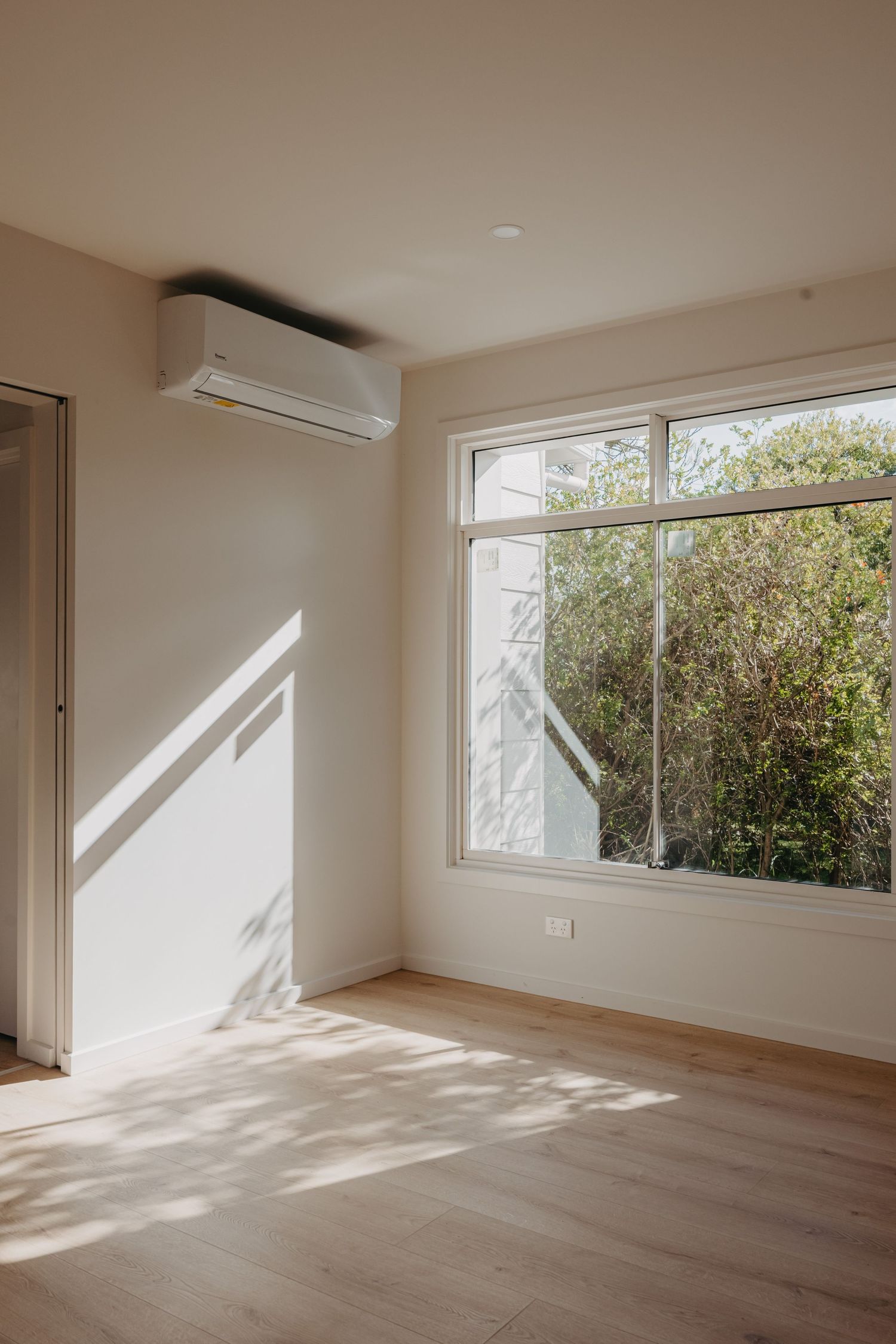 Bright minimalist bedroom with large window overlooking garden, light timber floors, and wall-mounted split system AC.