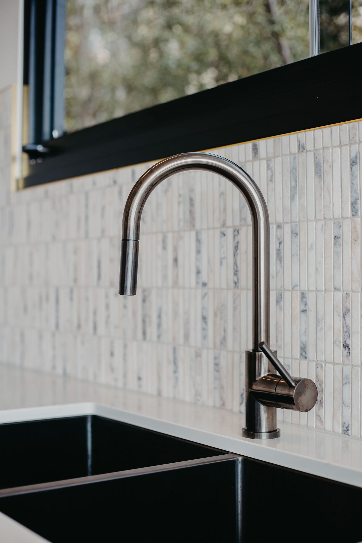 Brushed gunmetal gooseneck kitchen faucet over black sink with marble tile backsplash and black window frame.