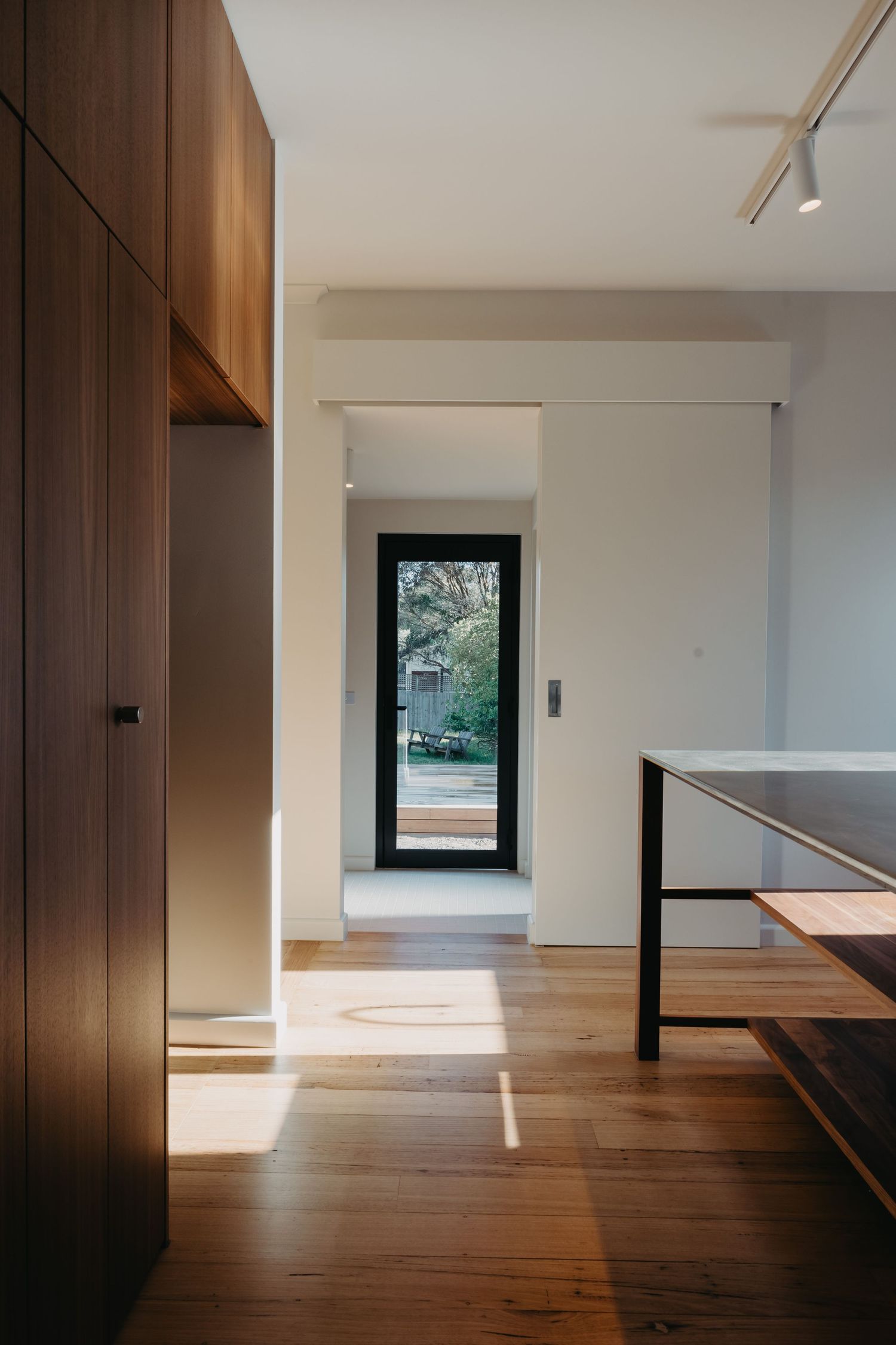 Sunlit interior hallway with walnut joinery, timber floors and black-framed glass door opening to outdoor timber deck.