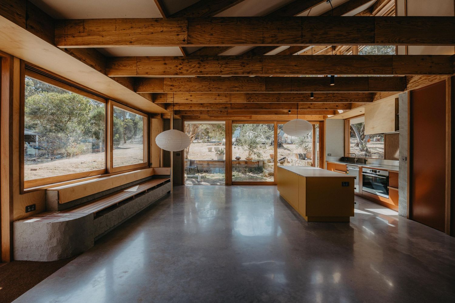 Open-plan living space with exposed timber beams, polished concrete floors, yellow kitchen island, and large windows.