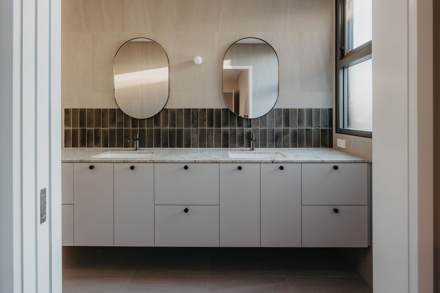 Modern bathroom with double oval mirrors, white floating vanity, marble countertop, and dark tile backsplash near a window.
