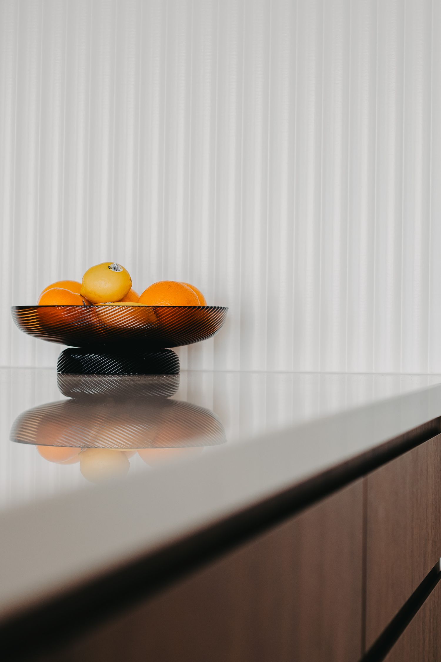 Close-up of oranges in a dark glass bowl on a white reflective countertop against a textured white wall panel.