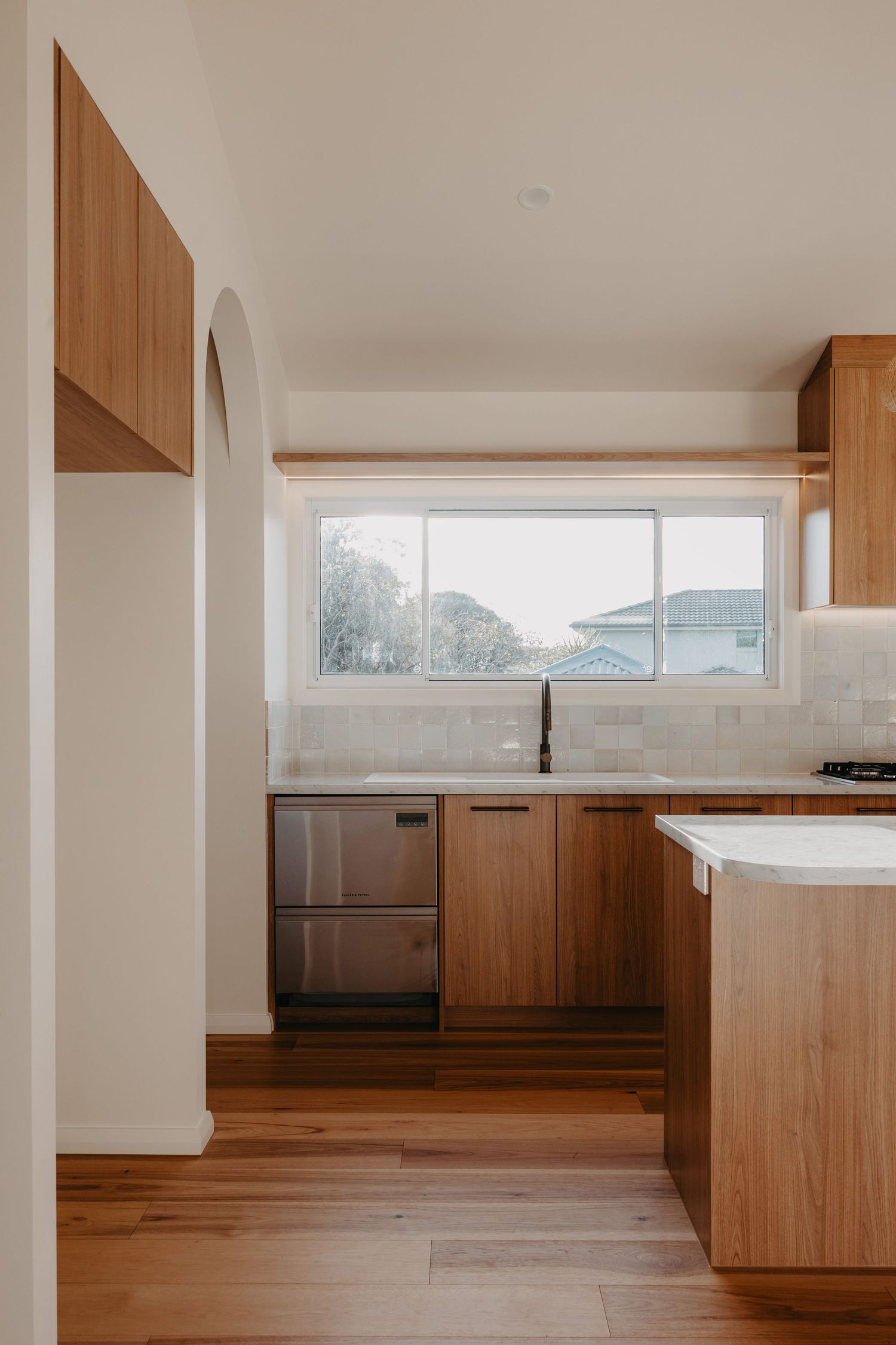 Spacious kitchen with timber cabinets, white stone island bench, arched alcove, large window, and light hardwood timber flooring.