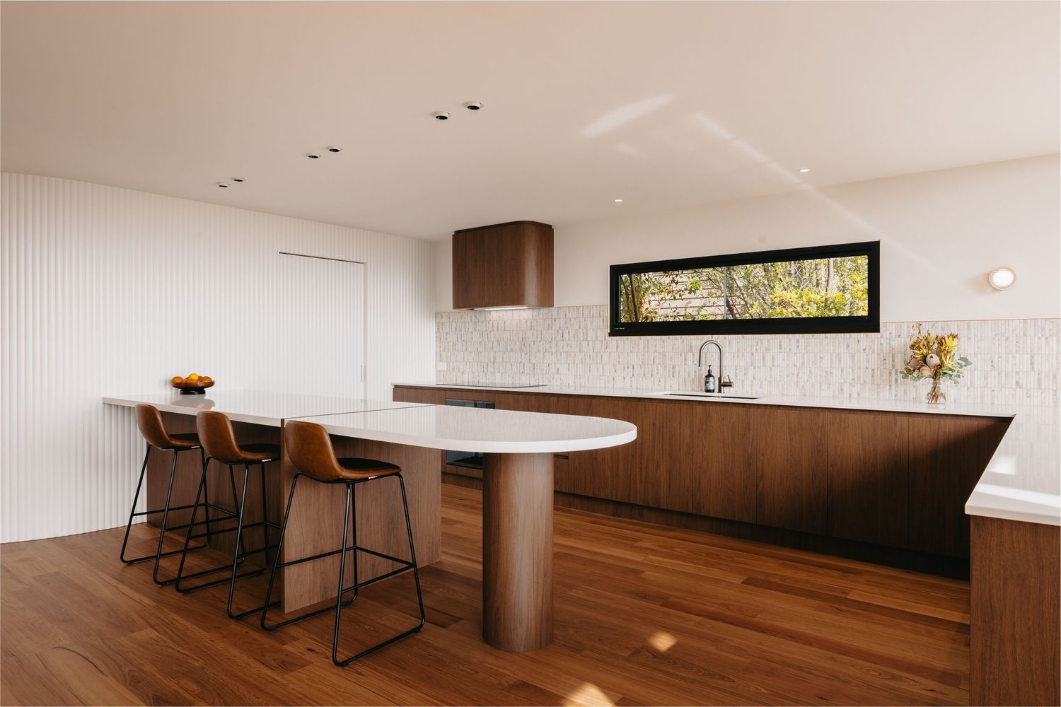 Bright modern kitchen with dark walnut cabinetry, oval island, leather bar stools, and horizontal window.
