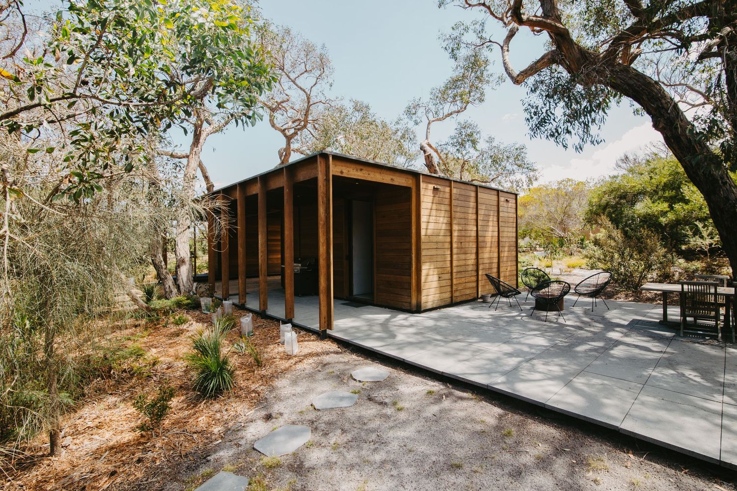 Modern wooden cabin with cedar siding nestled among oak trees on a stone patio in a natural landscape.
