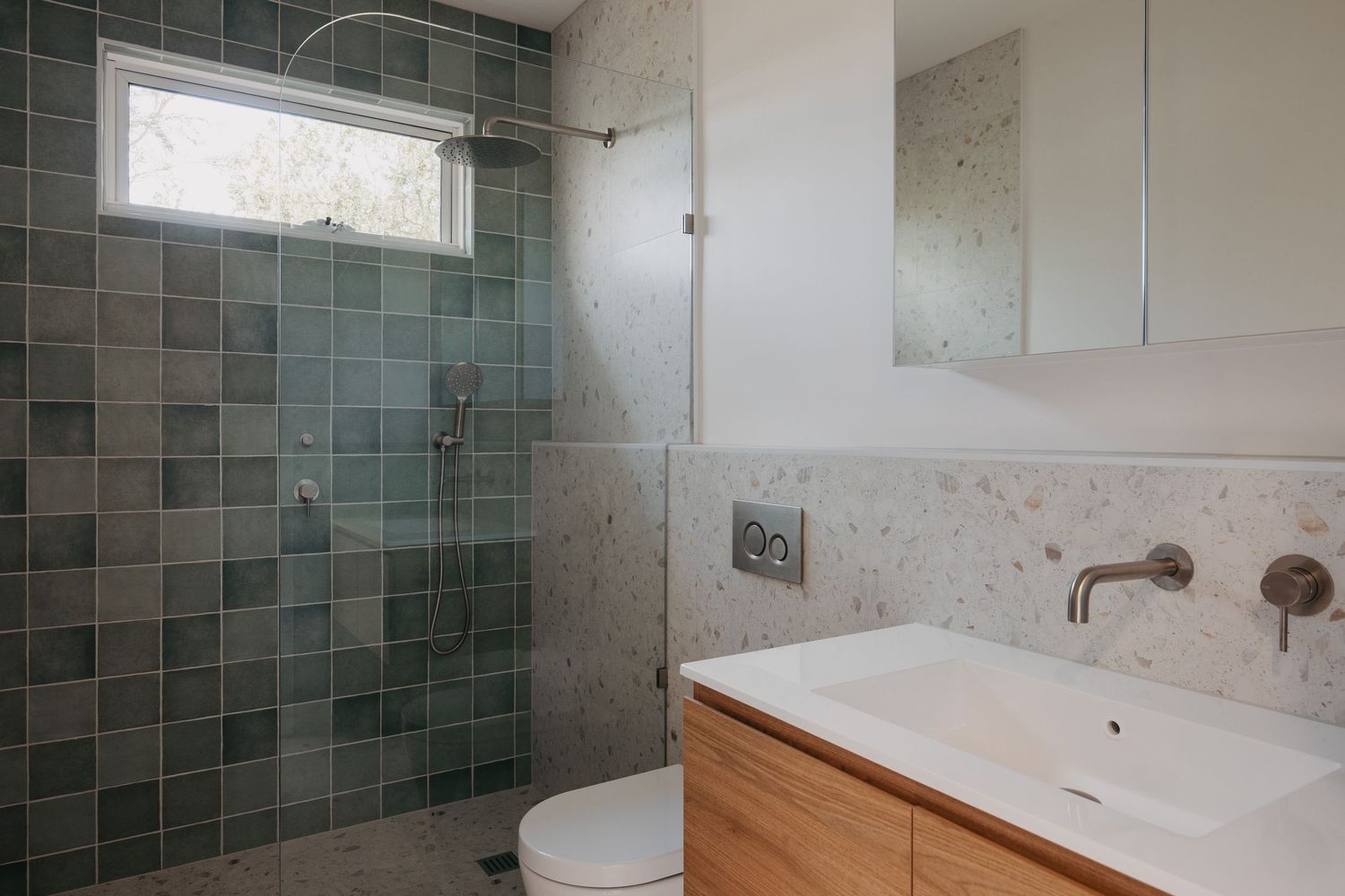 Modern bathroom with green tiled walk-in shower, terrazzo walls, wooden vanity, and white sink under a large mirror.