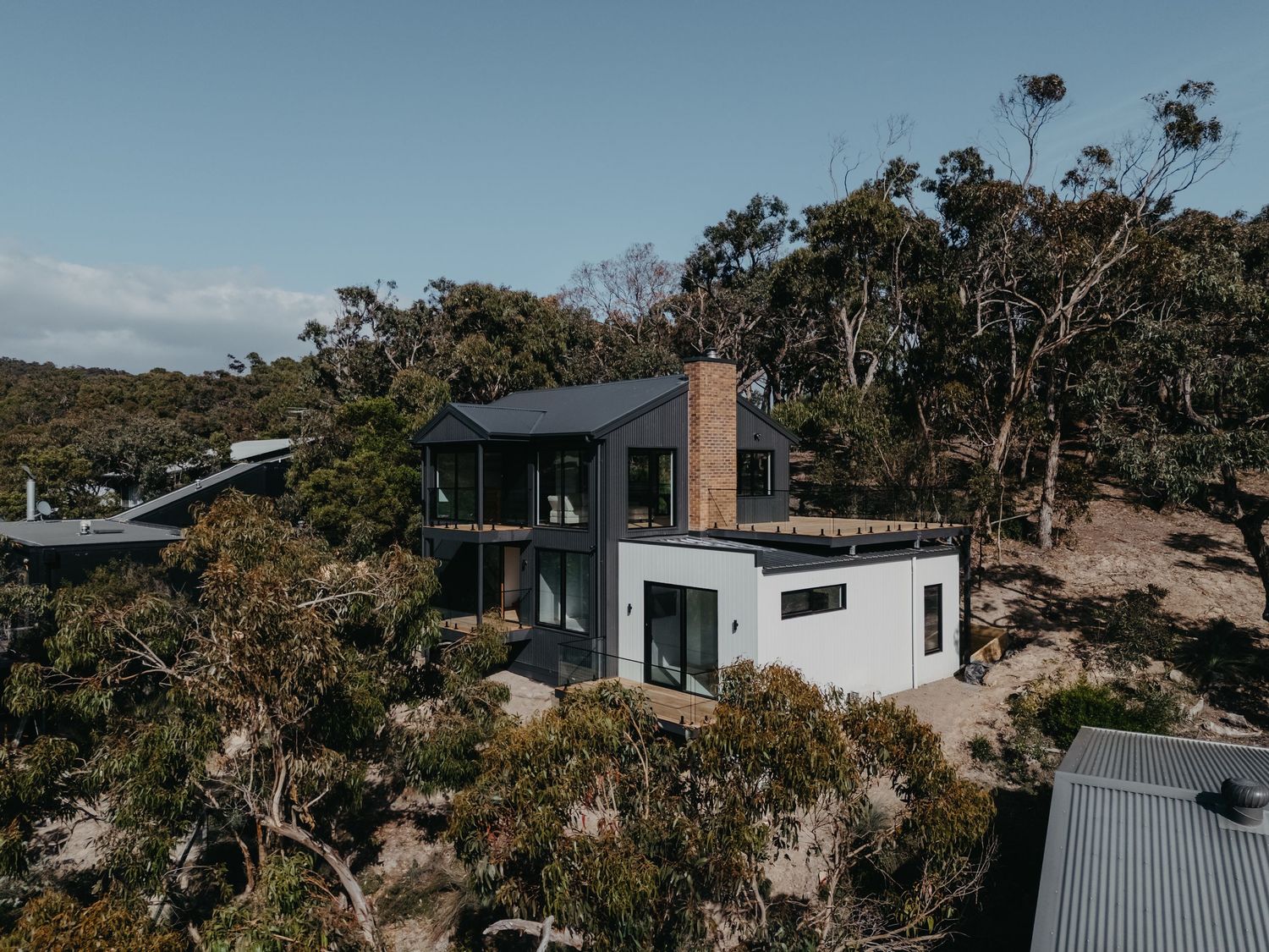 Aerial view of a modern two-storey home with dark roof and timber accents nestled among dense native bushland.