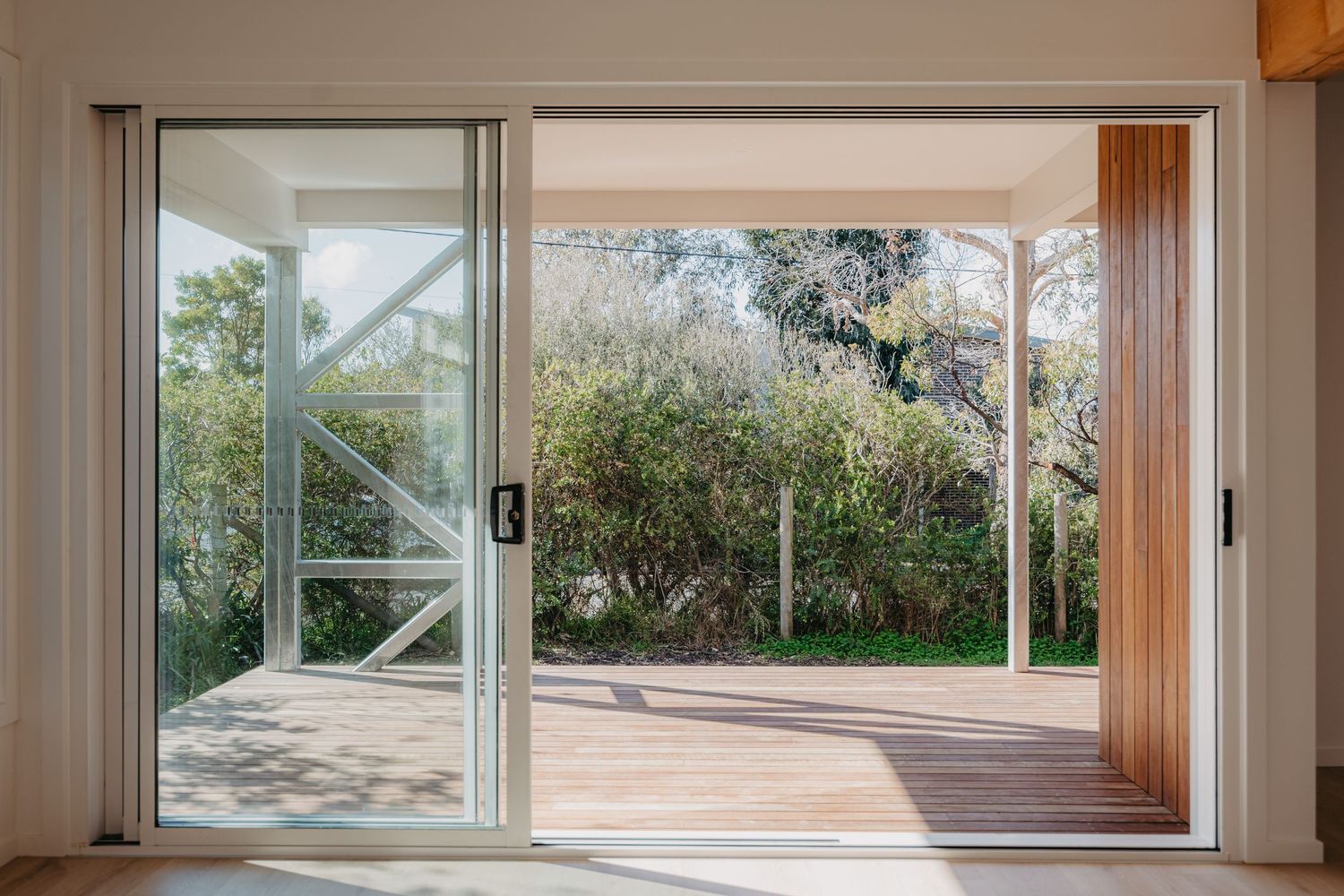 Open sliding glass doors leading to timber deck with lush green garden and steel frame pergola.