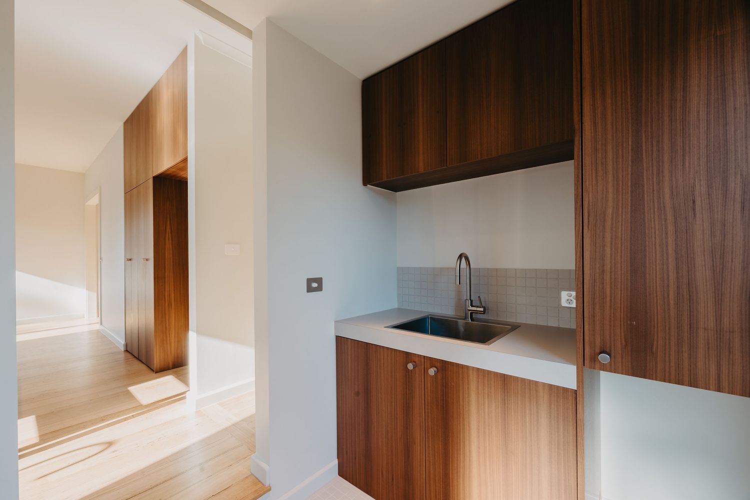 Modern laundry nook with dark walnut cabinetry, stainless steel sink, and light hardwood floors leading to hallway.