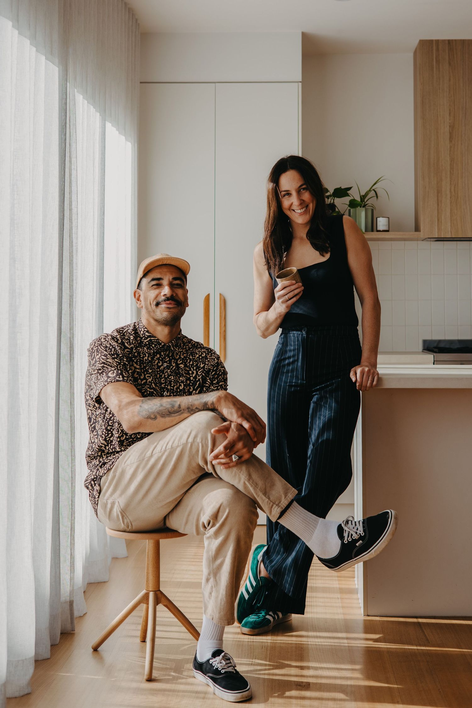 A smiling couple poses casually in a modern kitchen, man seated on stool, woman leaning on counter holding a drink.