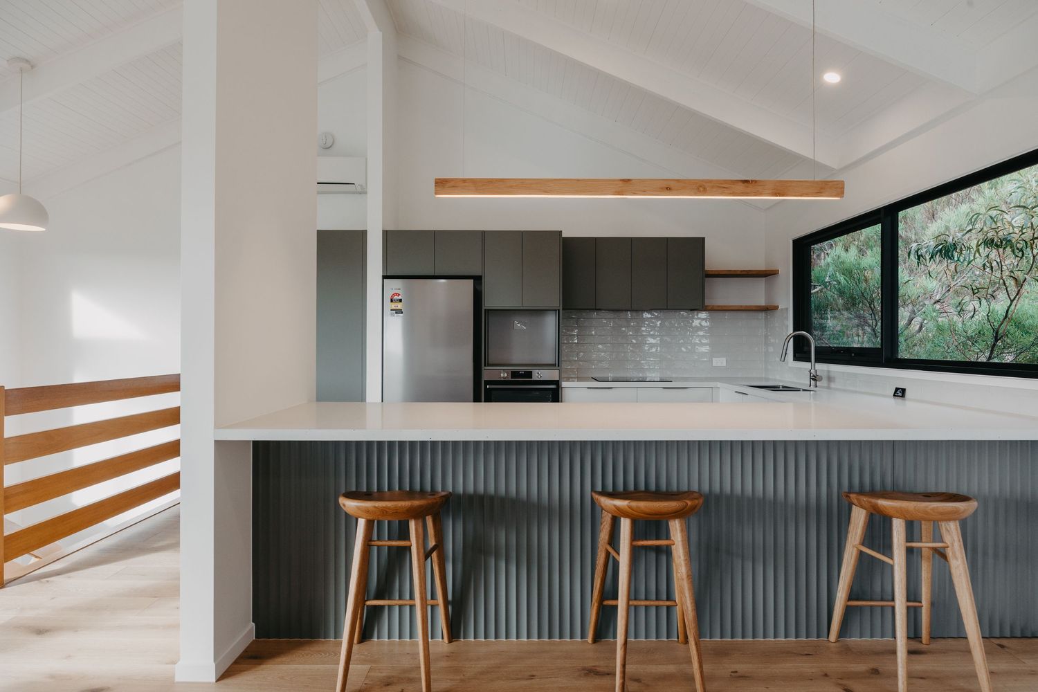 Modern kitchen with grey cabinetry, corrugated island bench, timber bar stools, and stainless steel fridge.