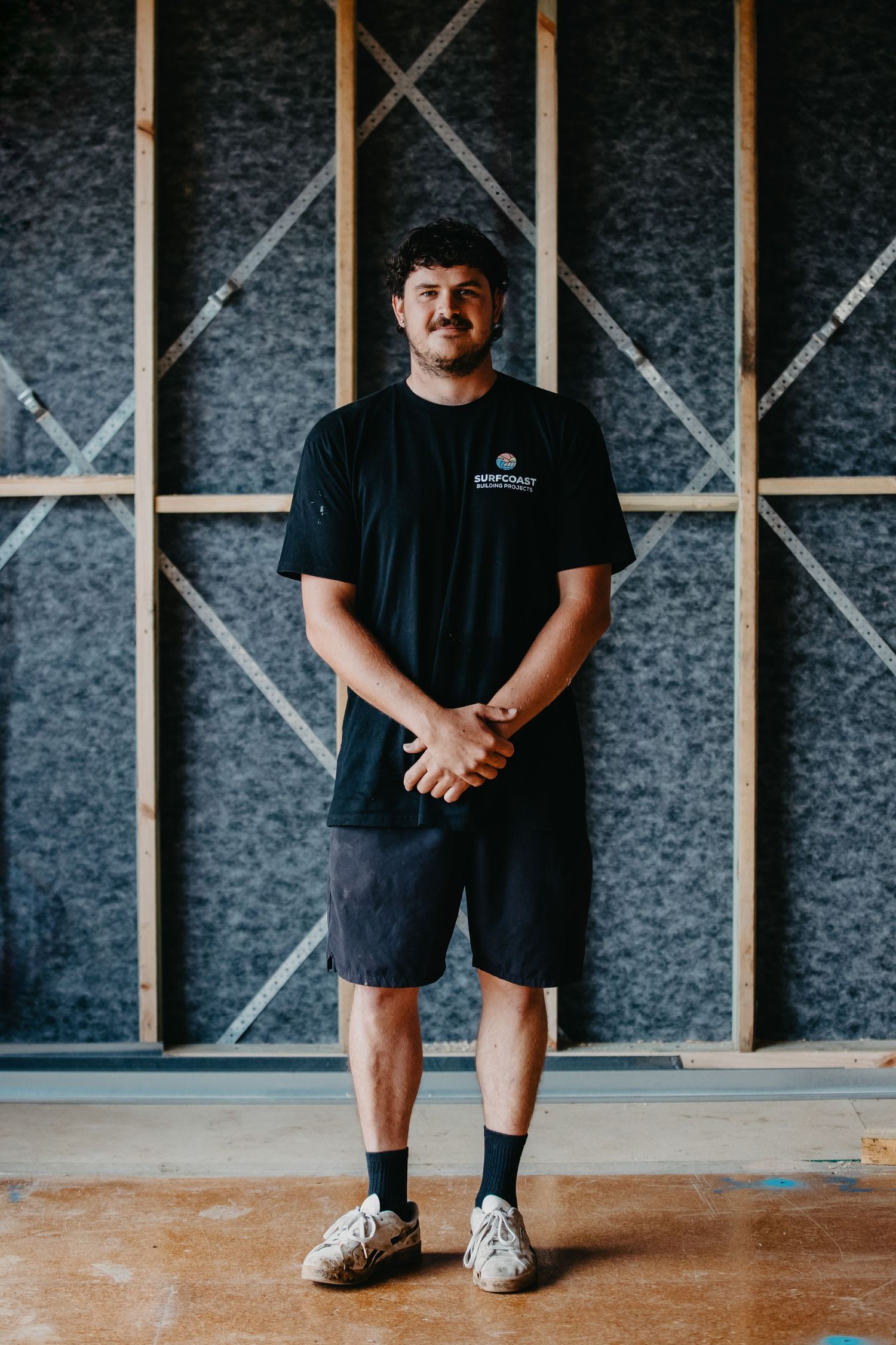 Young man in black Surfcoast t-shirt and shorts standing with hands clasped in a construction site.