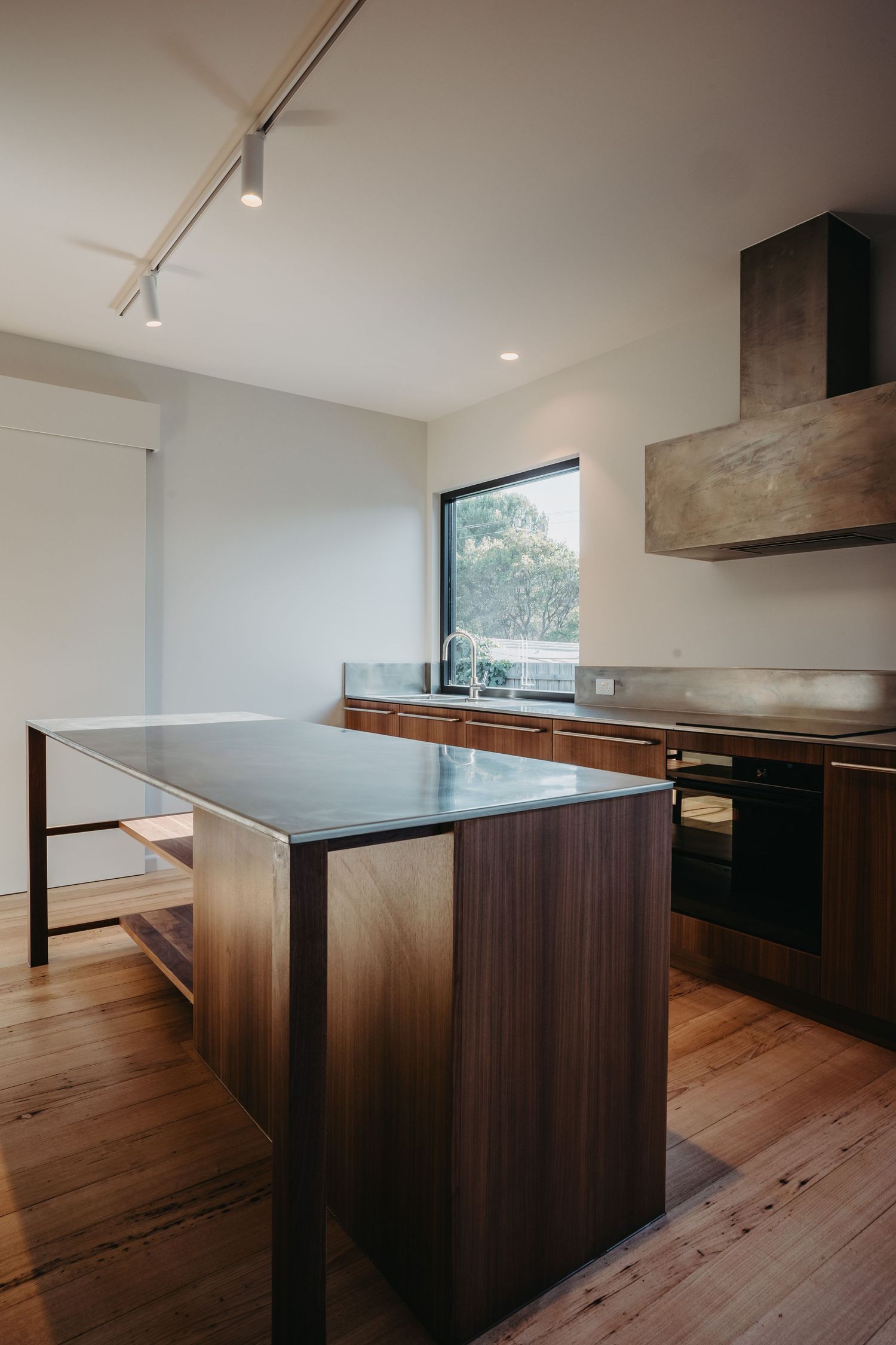 Dark walnut kitchen with glass-topped island bench, integrated appliances, range hood and black-framed window with garden views.