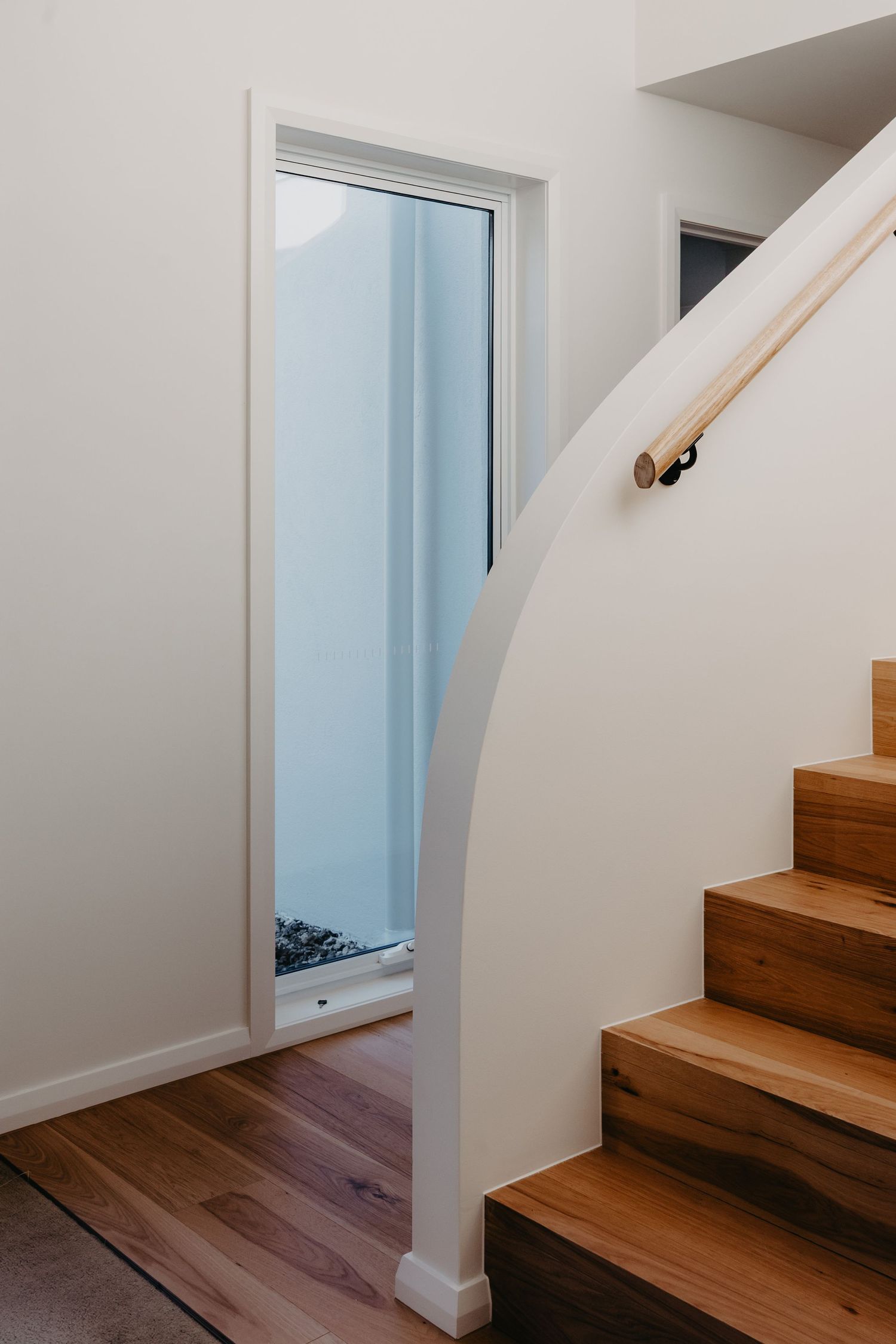 Modern staircase with timber steps, curved white balustrade, wooden handrail, and a tall narrow window flooding natural light.