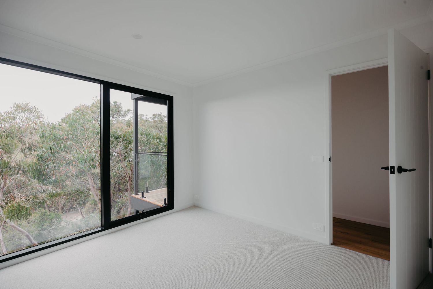Bright empty bedroom with black-framed floor-to-ceiling window overlooking lush green treetop views.