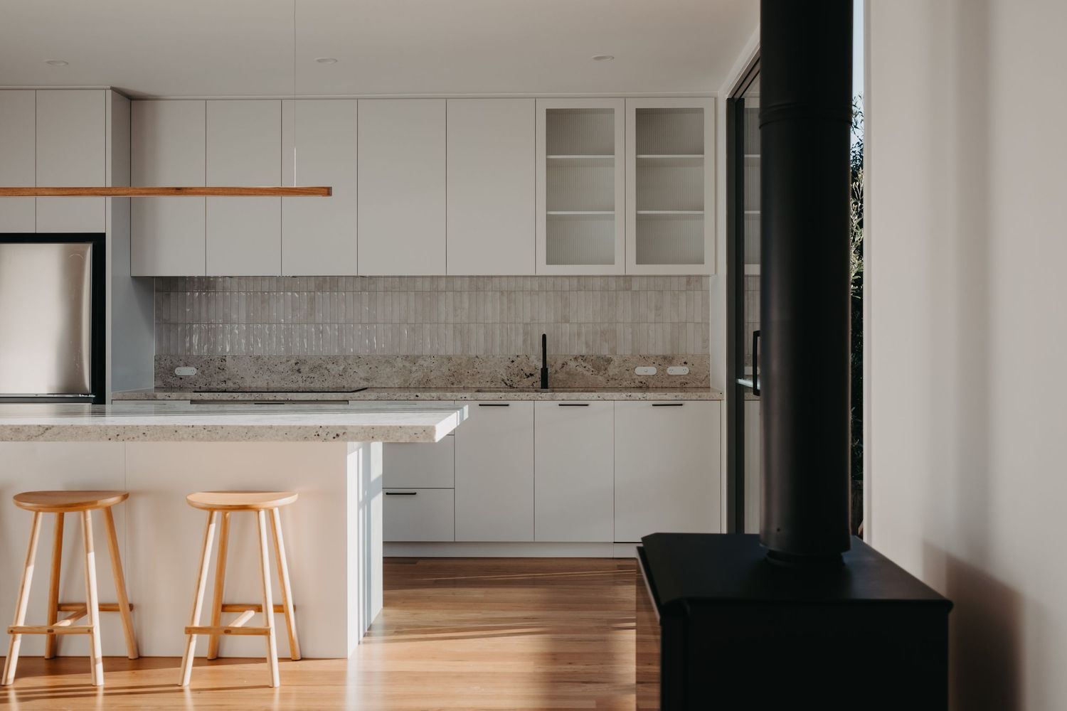 Modern minimalist kitchen with white cabinets, herringbone tile backsplash, wooden bar stools, and a black wood-burning stove.