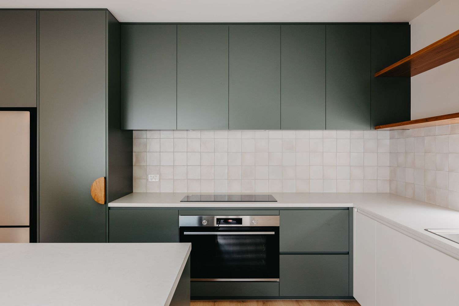 Modern kitchen with dark green cabinetry, white tile backsplash, built-in oven, and sleek countertops.