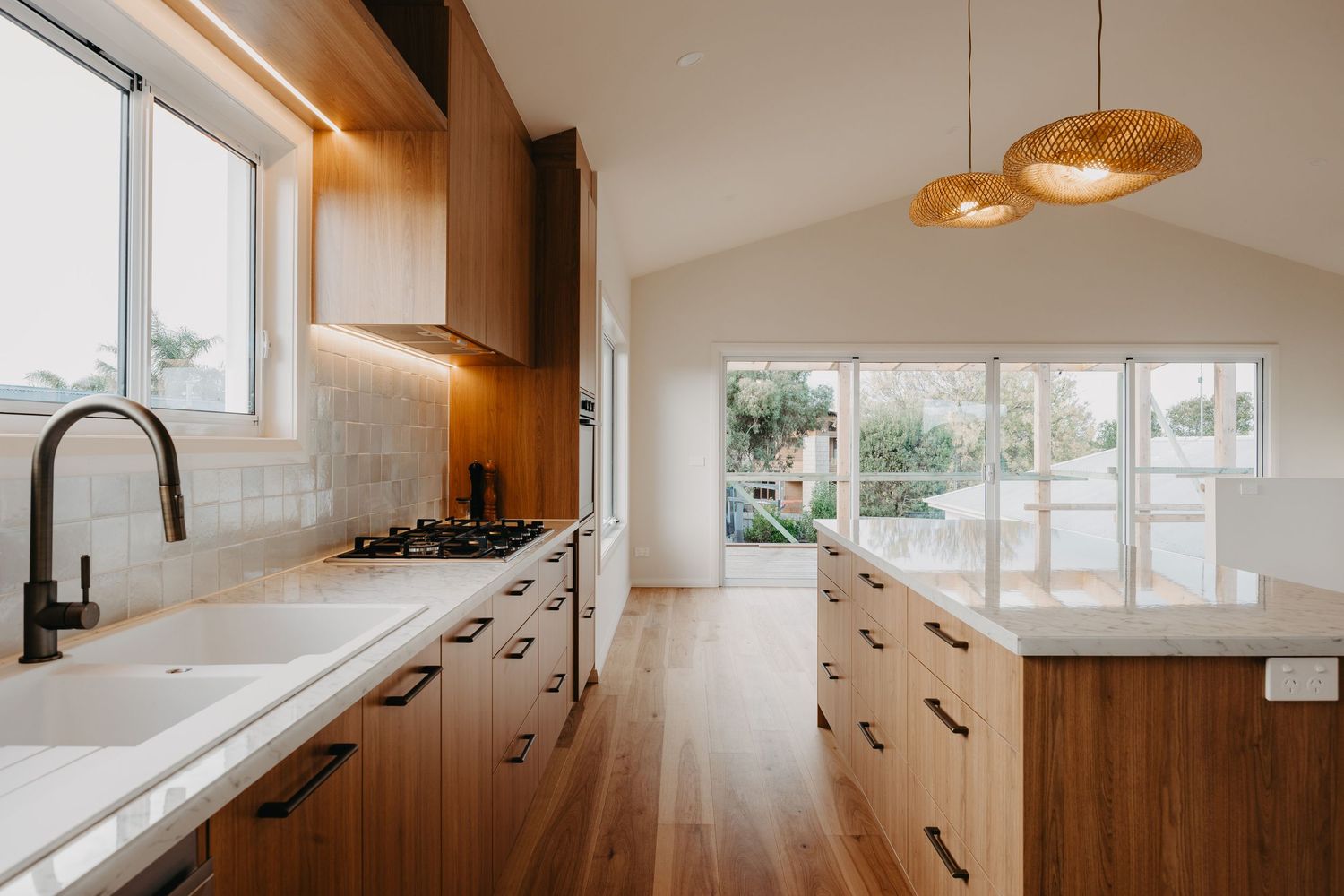 Bright modern kitchen with timber cabinetry, white stone benchtops, black tapware, gas cooktop, and large windows overlooking outdoor area.