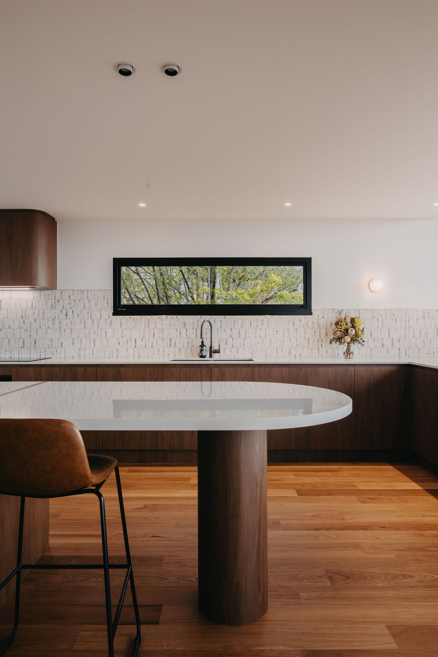 Modern kitchen with walnut cabinets, white island, leather barstool, hardwood floors, and a horizontal window above the sink.