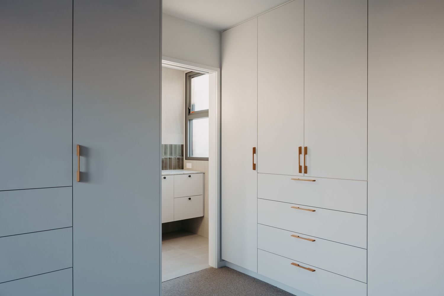Modern built-in wardrobe with white cabinetry and gold handles, adjacent to a bright ensuite bathroom.