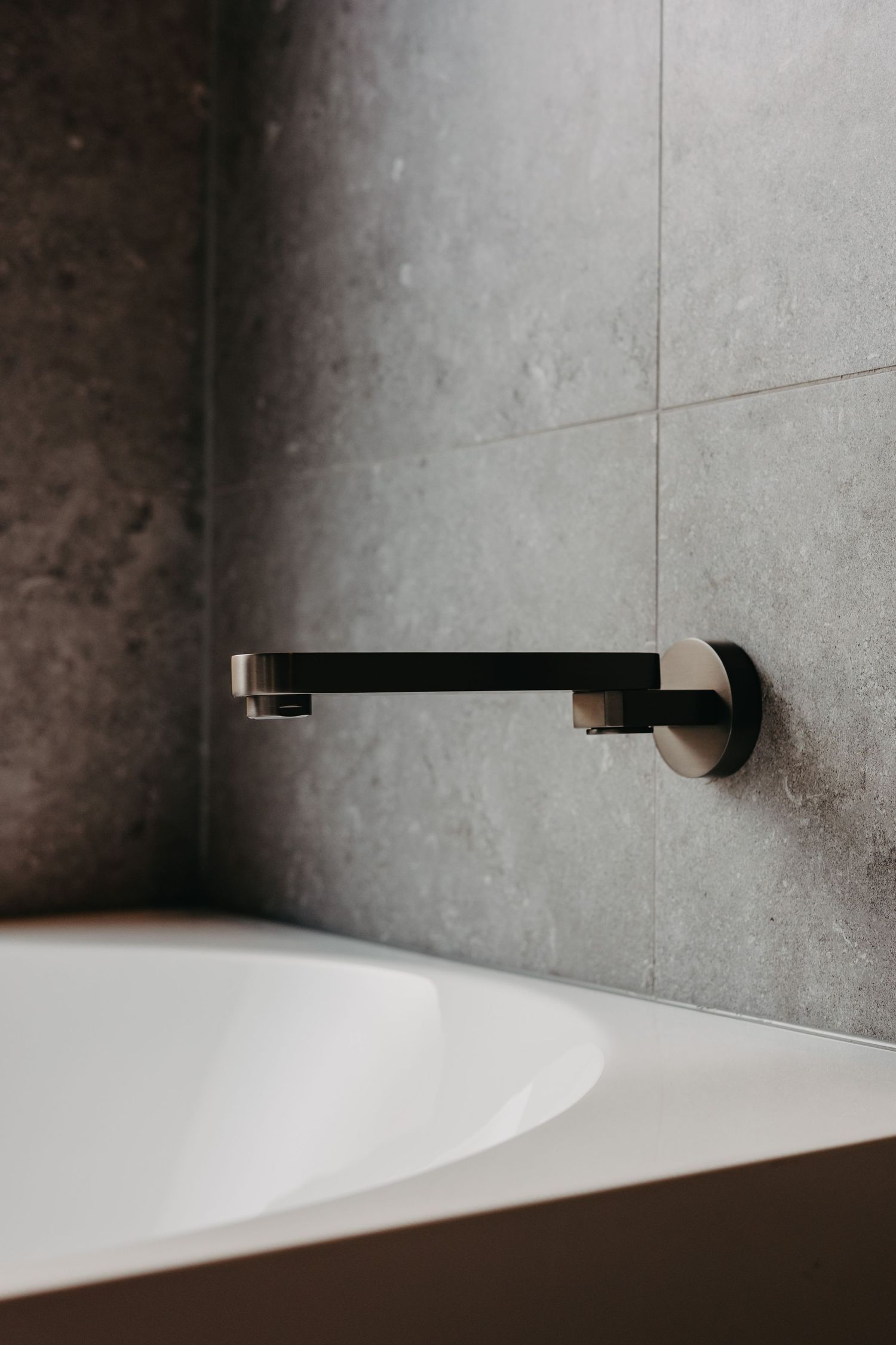 Minimalist bathroom with a matte black wall-mounted bath spout, grey stone tiles, and a white freestanding bathtub.
