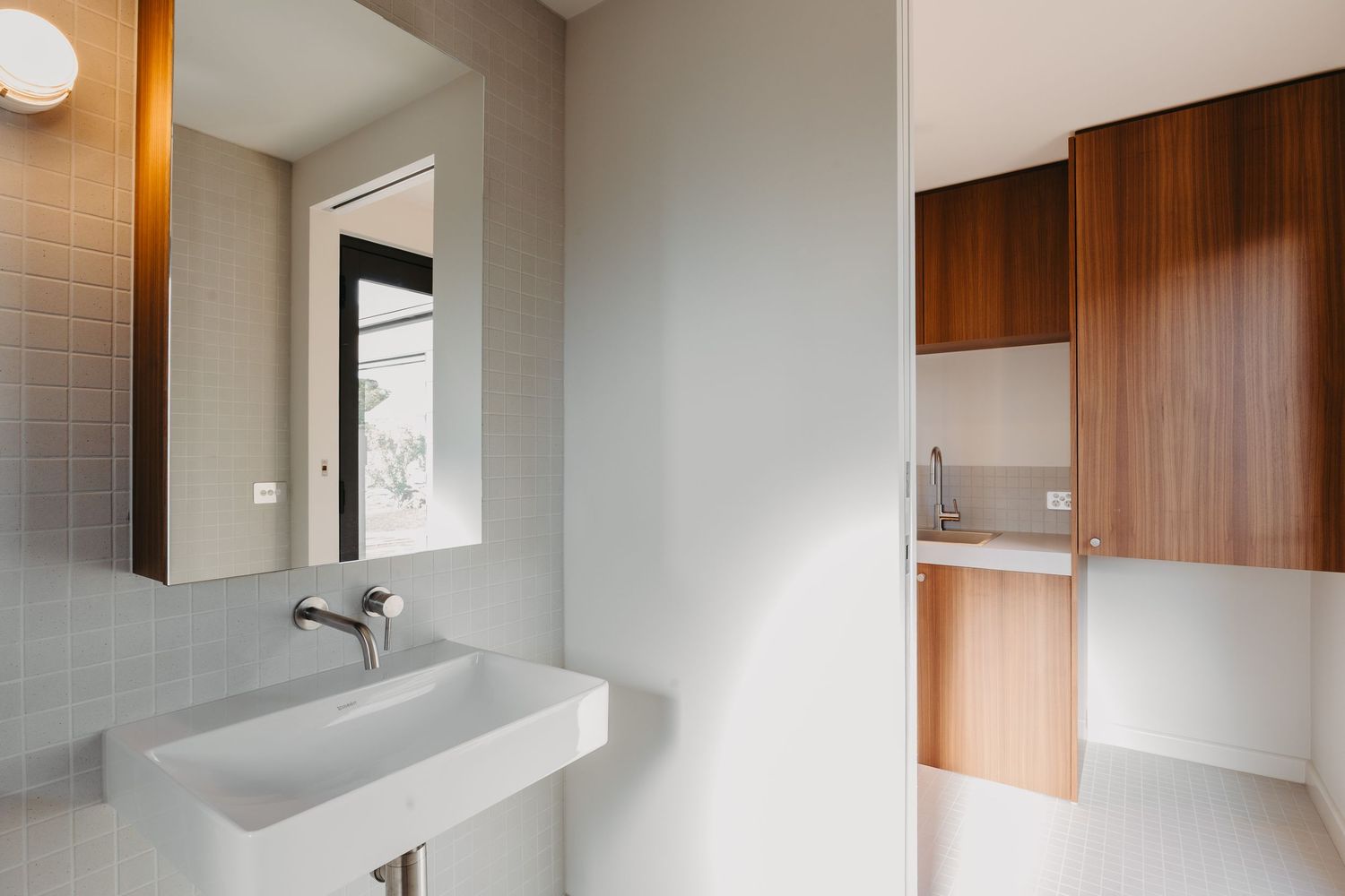 Modern bathroom with wall-mounted white basin, square mirror, mosaic tile walls and adjacent walnut storage cabinetry.