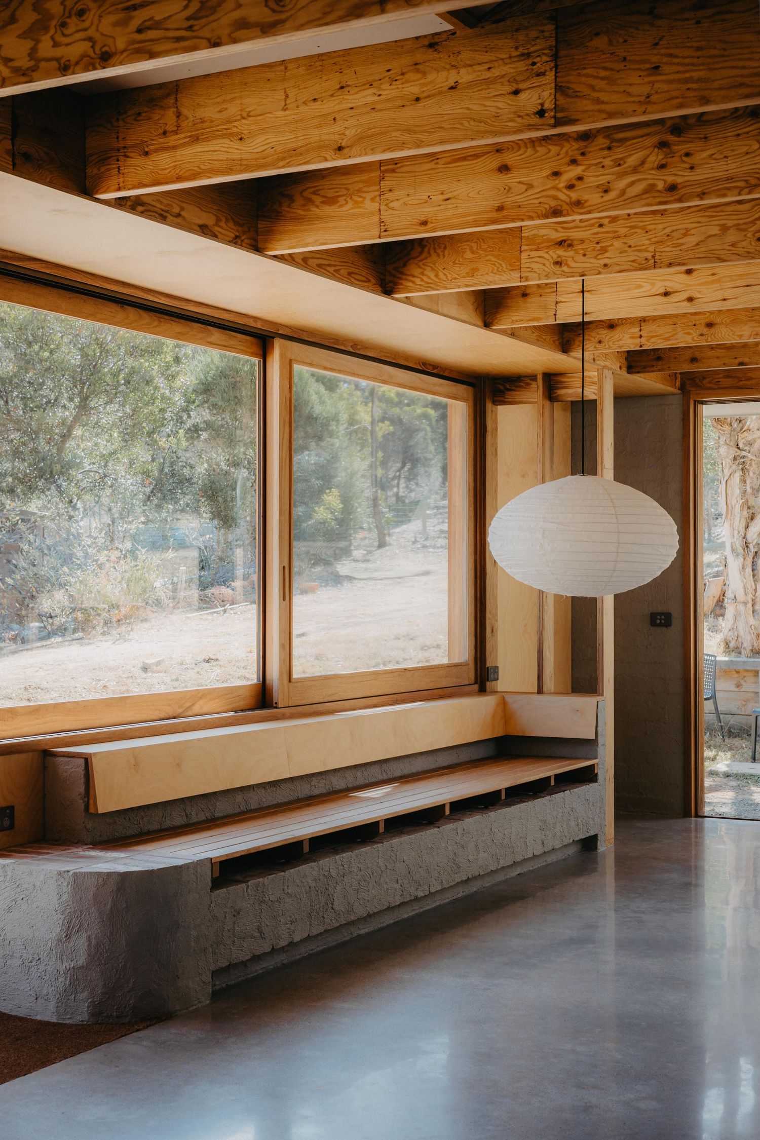 Timber-framed interior with concrete bench seat, large windows overlooking bushland, and paper lantern light.