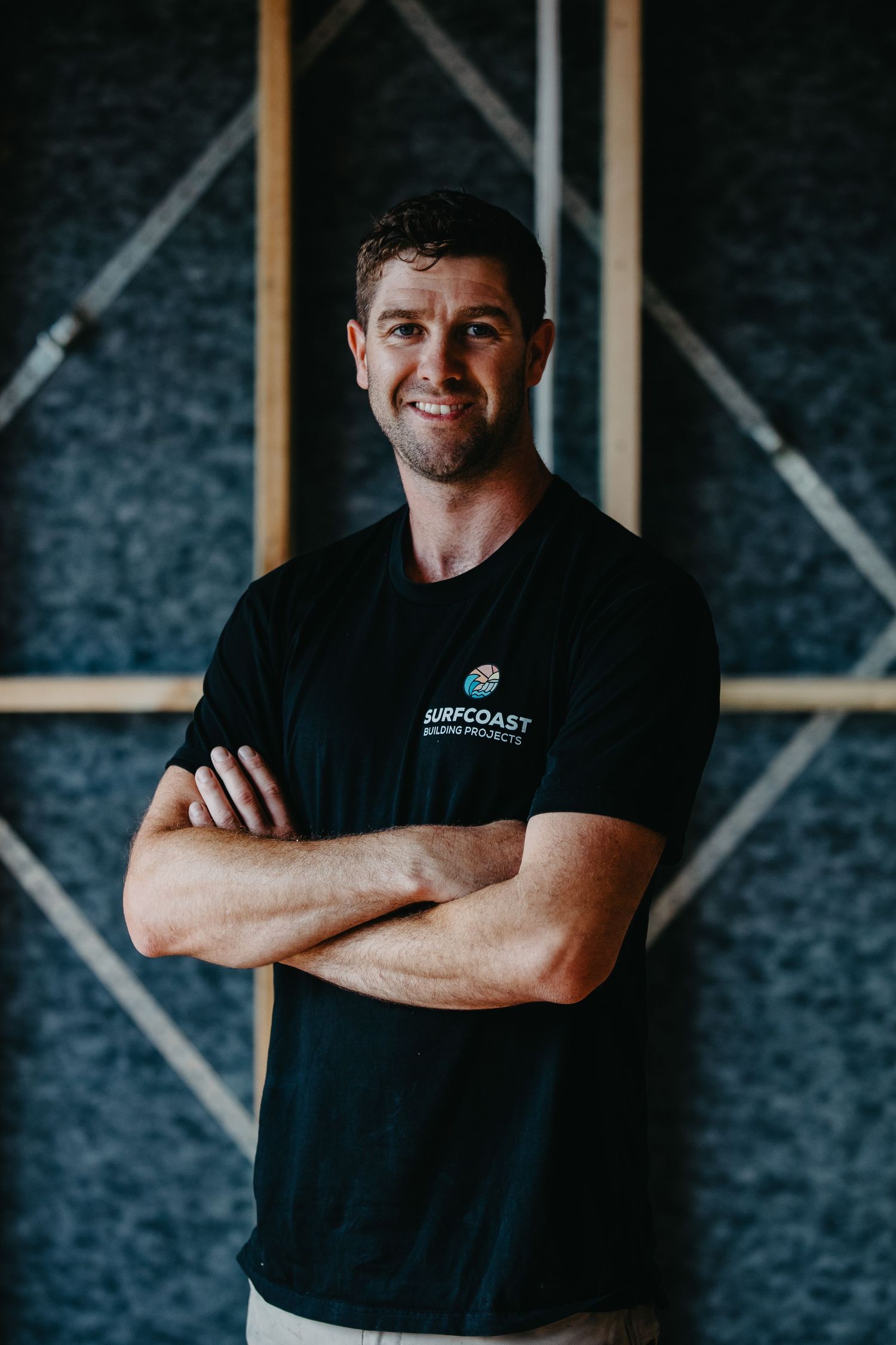 Smiling man in black Surfcoast t-shirt standing with arms crossed in a modern interior space.